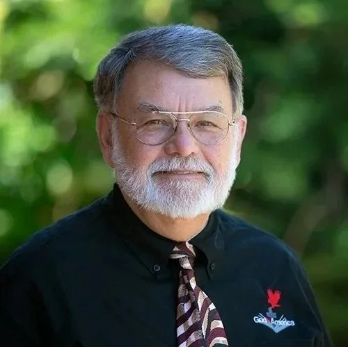 A portrait of an older man with glasses, gray hair, and a beard, wearing a black shirt with a logo, and a patterned tie, outdoors with a blurred green background.
