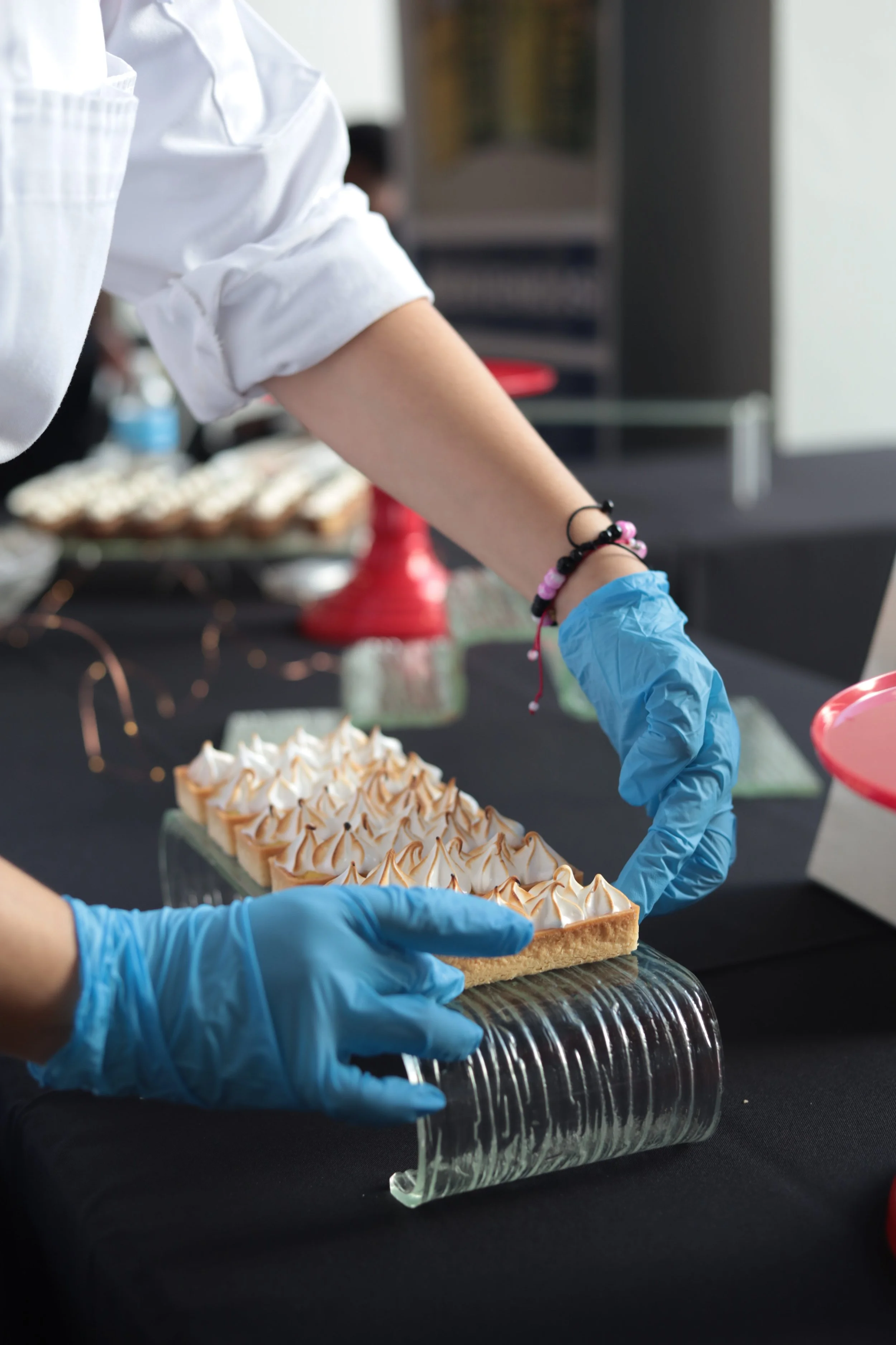 Person wearing blue gloves decorating a rectangular dessert with toasted meringue peaks at a black-covered table.