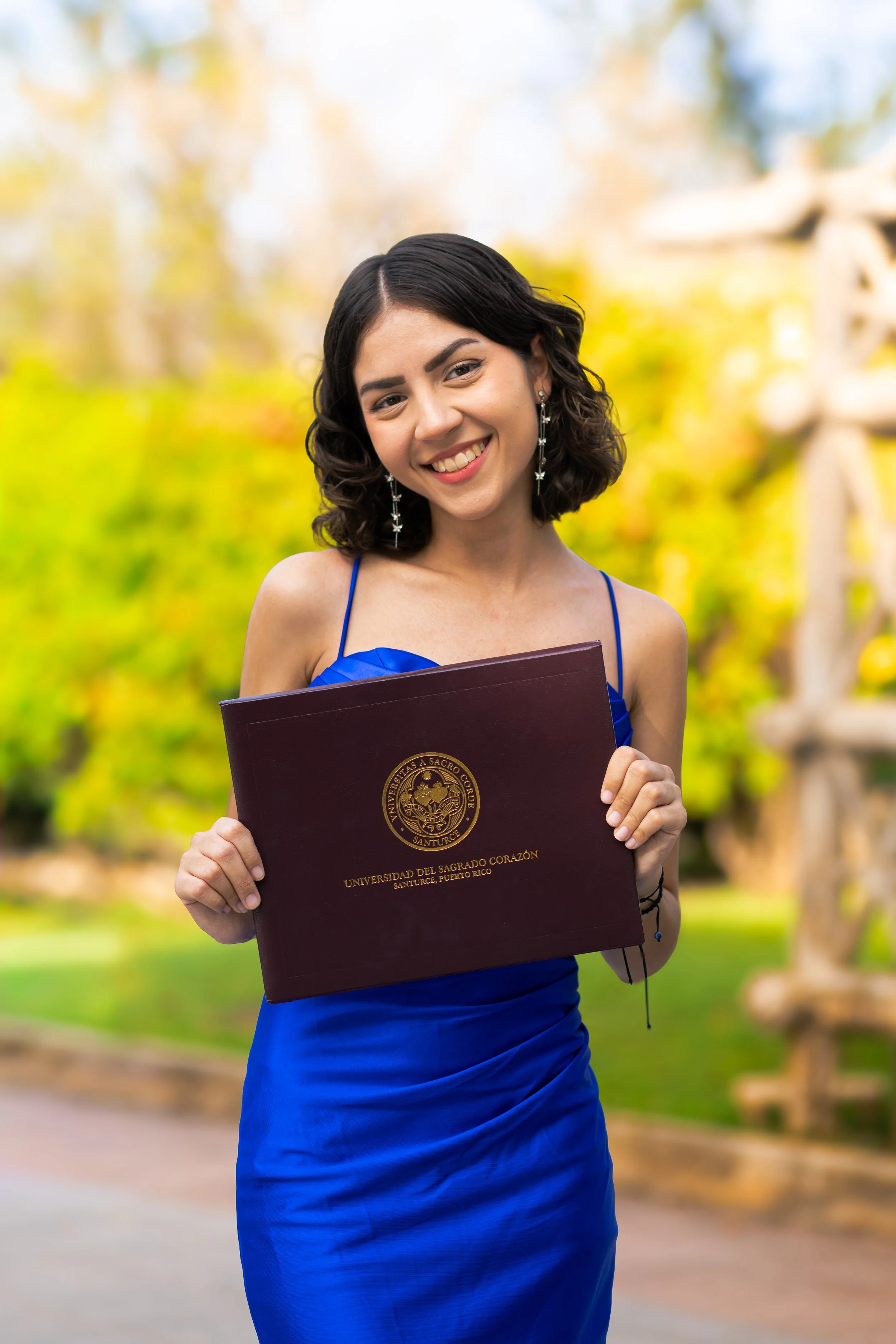 A young woman dressed in a blue gown, smiling and holding a diploma from Universidad del Sagrado Corazón, with a blurred outdoor background of trees and a sculpture.