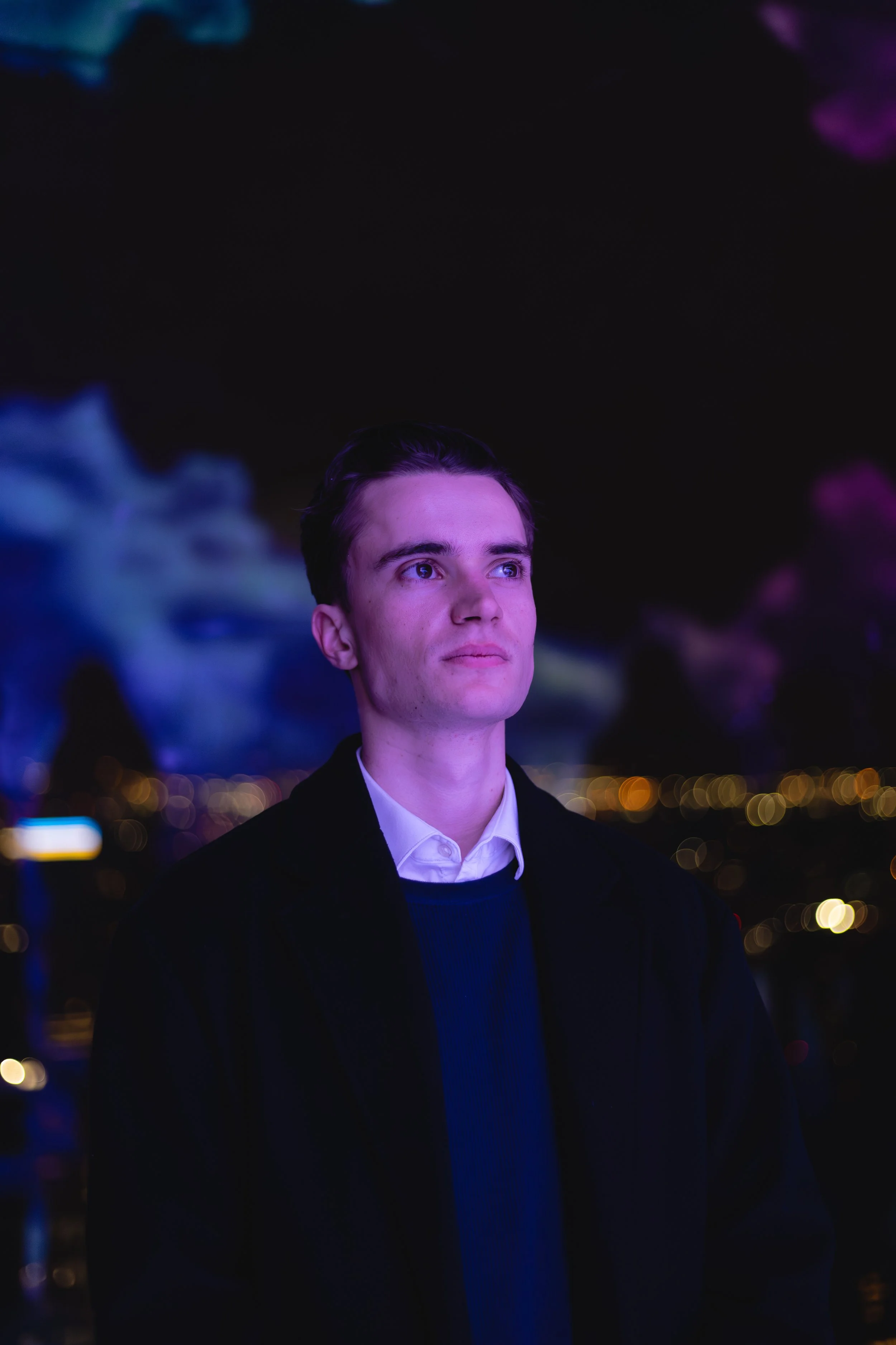 A young man with fair skin and dark hair standing outdoors at night with city lights and colorful smoke in the background.