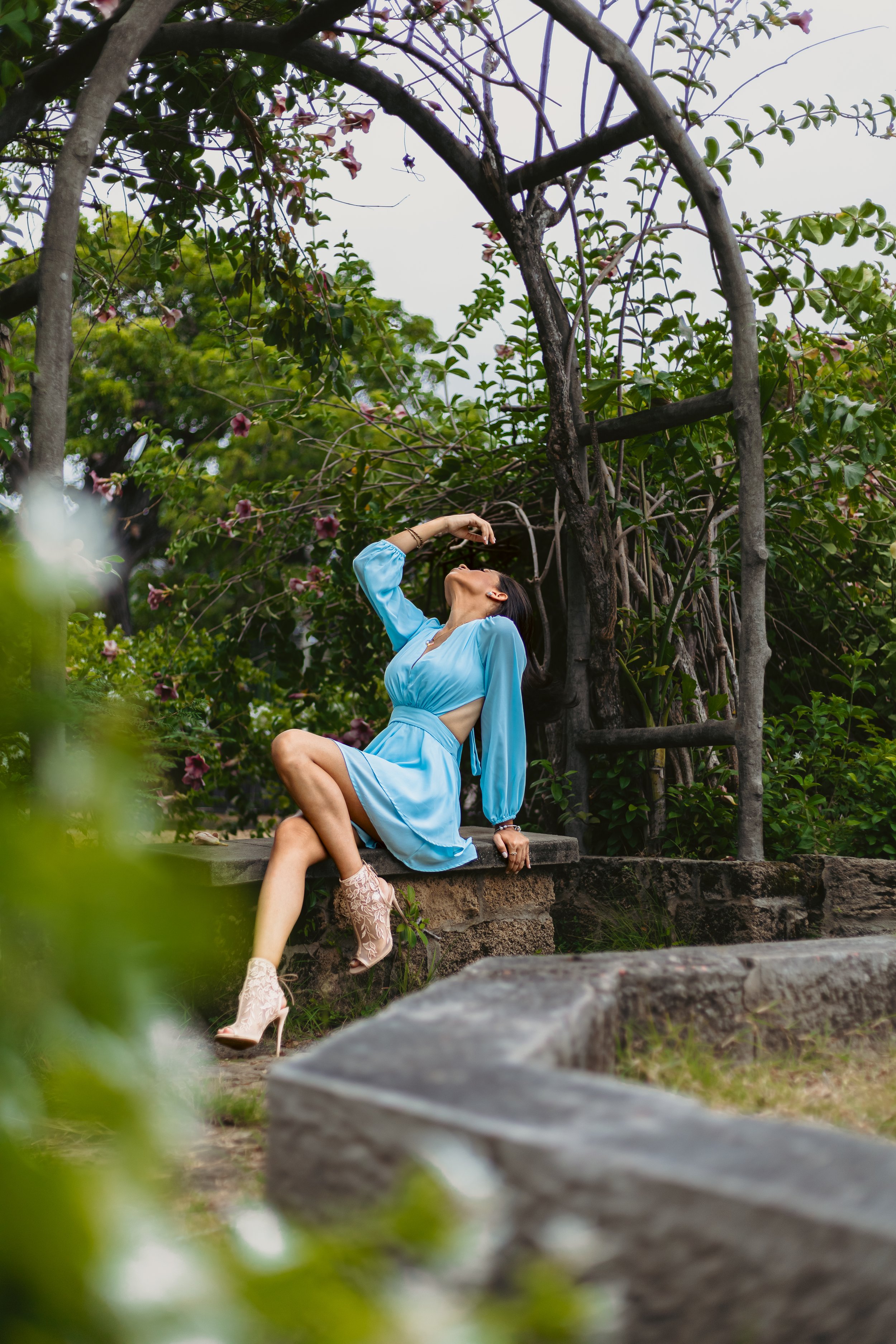 A woman in a blue dress and beige high heels sitting on a stone ledge outdoors, leaning back with her eyes closed and one hand on her forehead, surrounded by green trees and plants.
