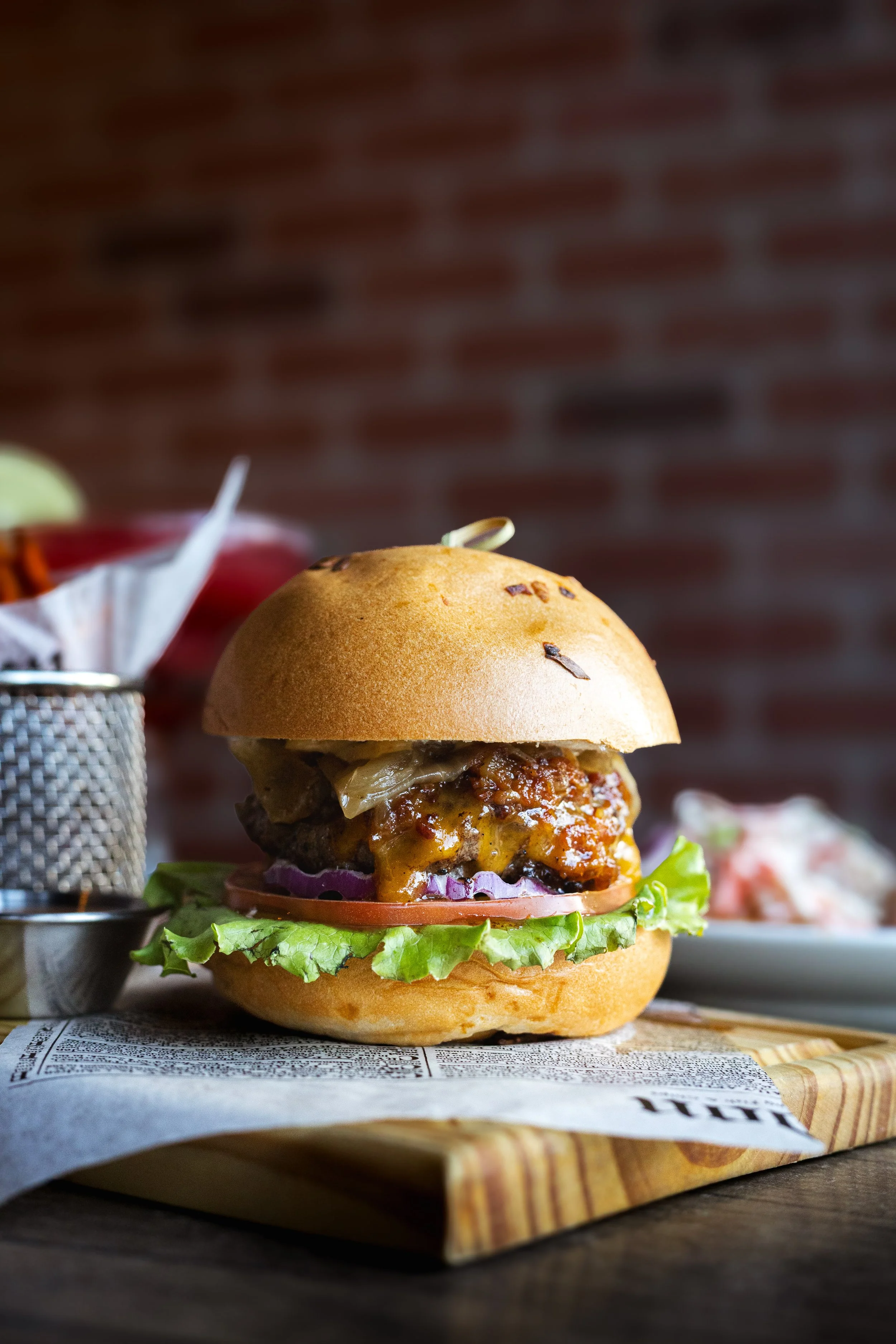 A close-up of a gourmet burger with lettuce, tomato, onions, cheese, a beef patty, and sauce in a toasted bun, placed on a wooden tray with a piece of newspaper underneath, against a blurred brick wall background.