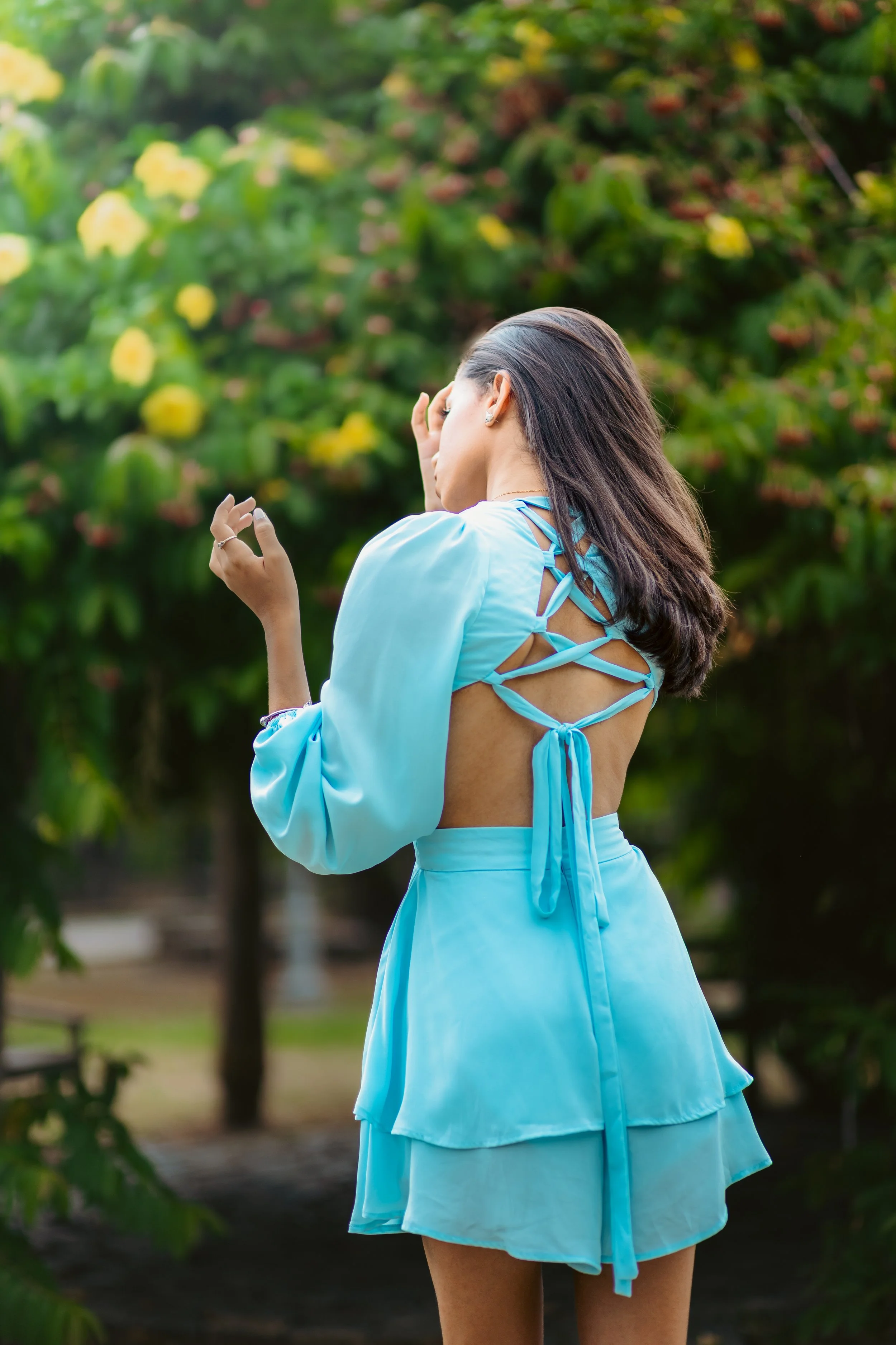 A woman in a blue dress with open back and tie details, standing outdoors near green trees with yellow flowers.
