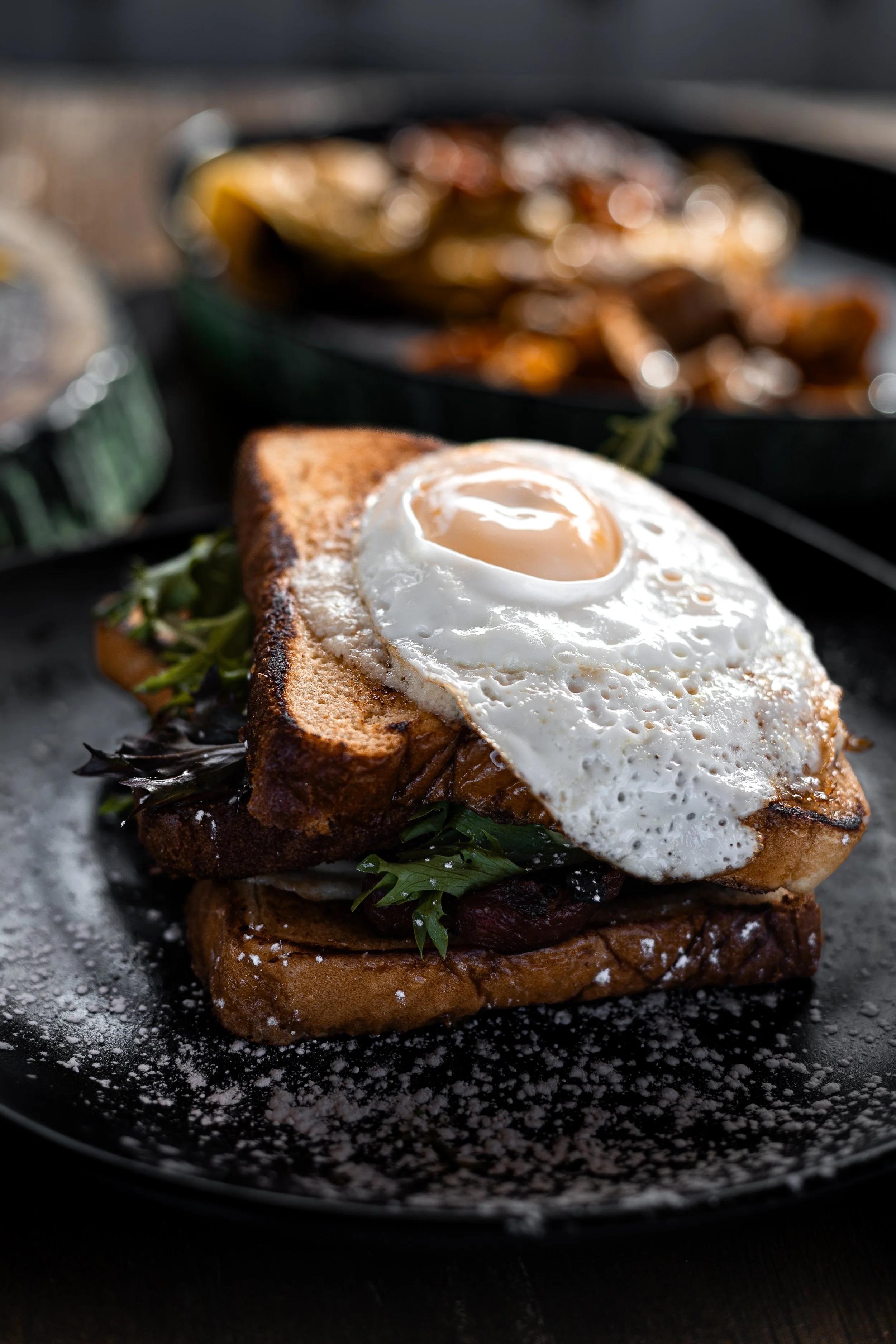 A gourmet breakfast sandwich with a fried egg on top, served on toasted bread, garnished with greens, and sprinkled with powdered sugar, on a black plate.