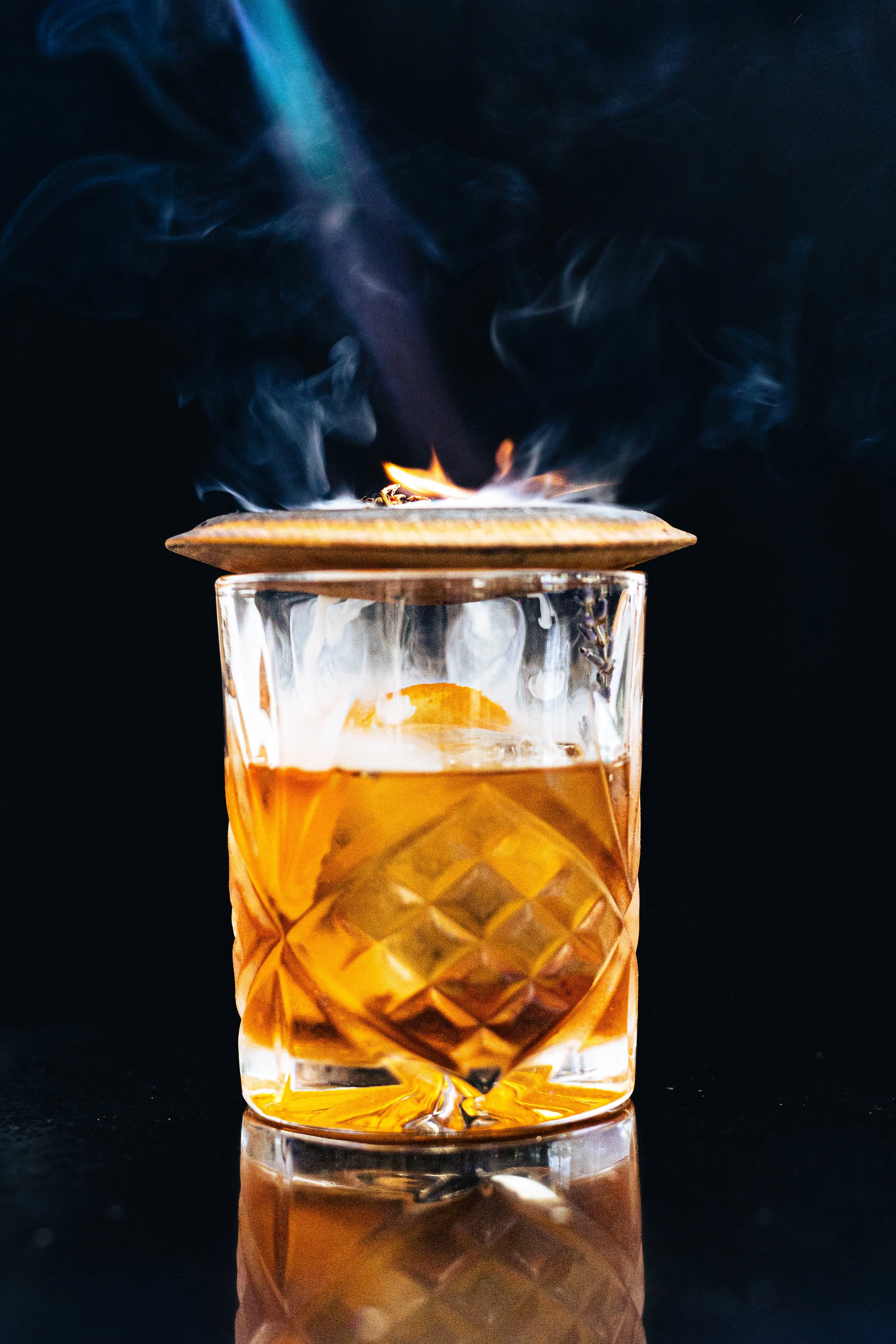A glass of whiskey with a large ice cube, topped with a wooden disc on which a small flame is burning, with swirling smoke in the background.