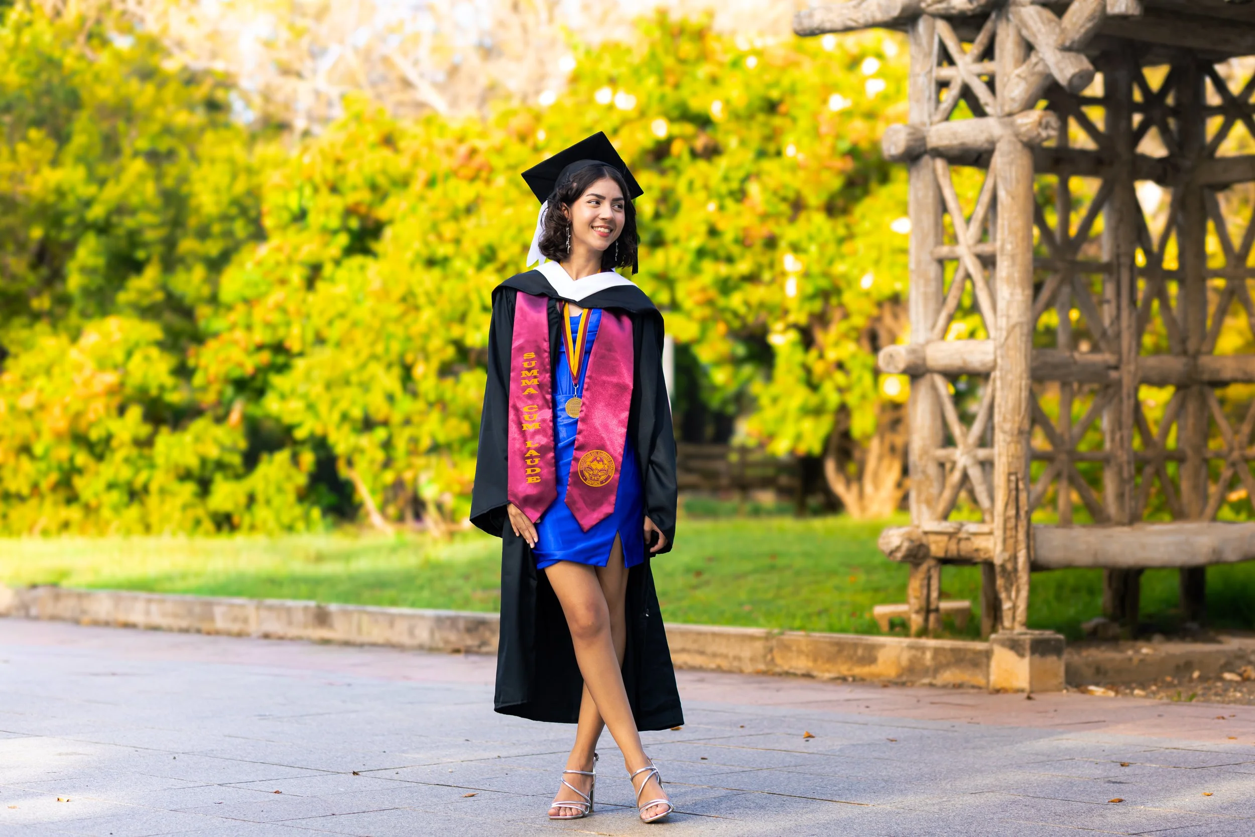 A woman in a graduation gown and cap smiling outdoors, wearing a pink and gold stole and medals, standing on a paved path with green trees and autumn foliage in the background.