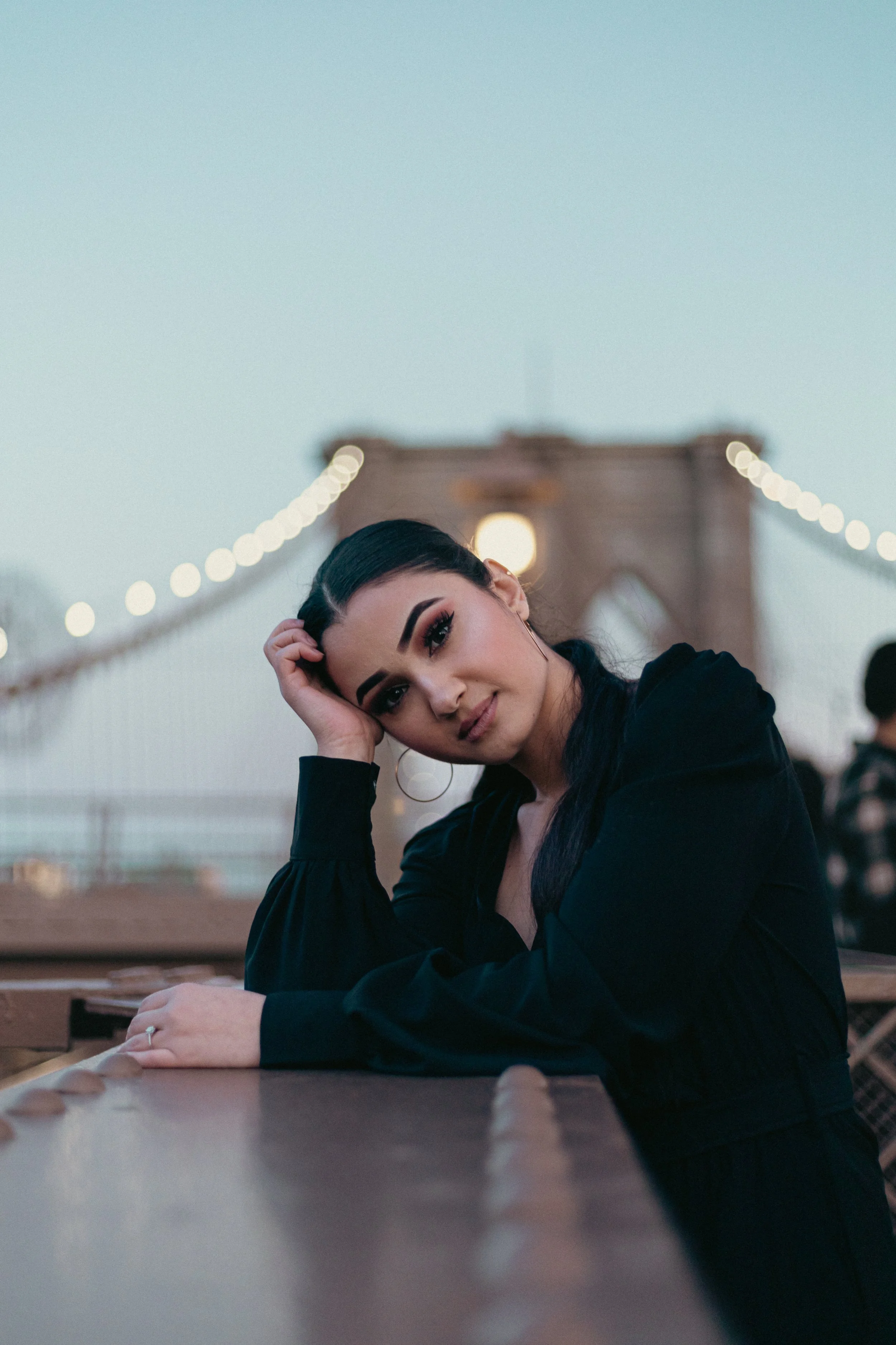 A woman sitting at a table on a bridge at dusk, resting her head on her hand, with string lights and the bridge structure in the background.