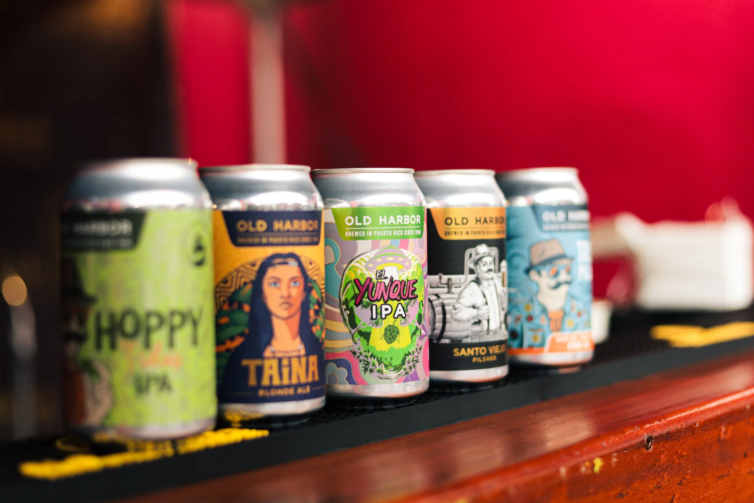A row of colorful craft beer cans on a bar counter with a blurred red background.