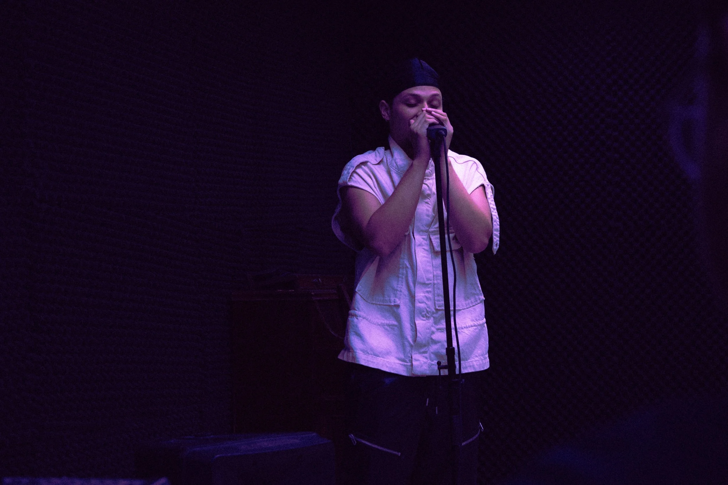 A woman singing into a microphone on stage, illuminated by purple lighting, with a dark textured wall in the background.