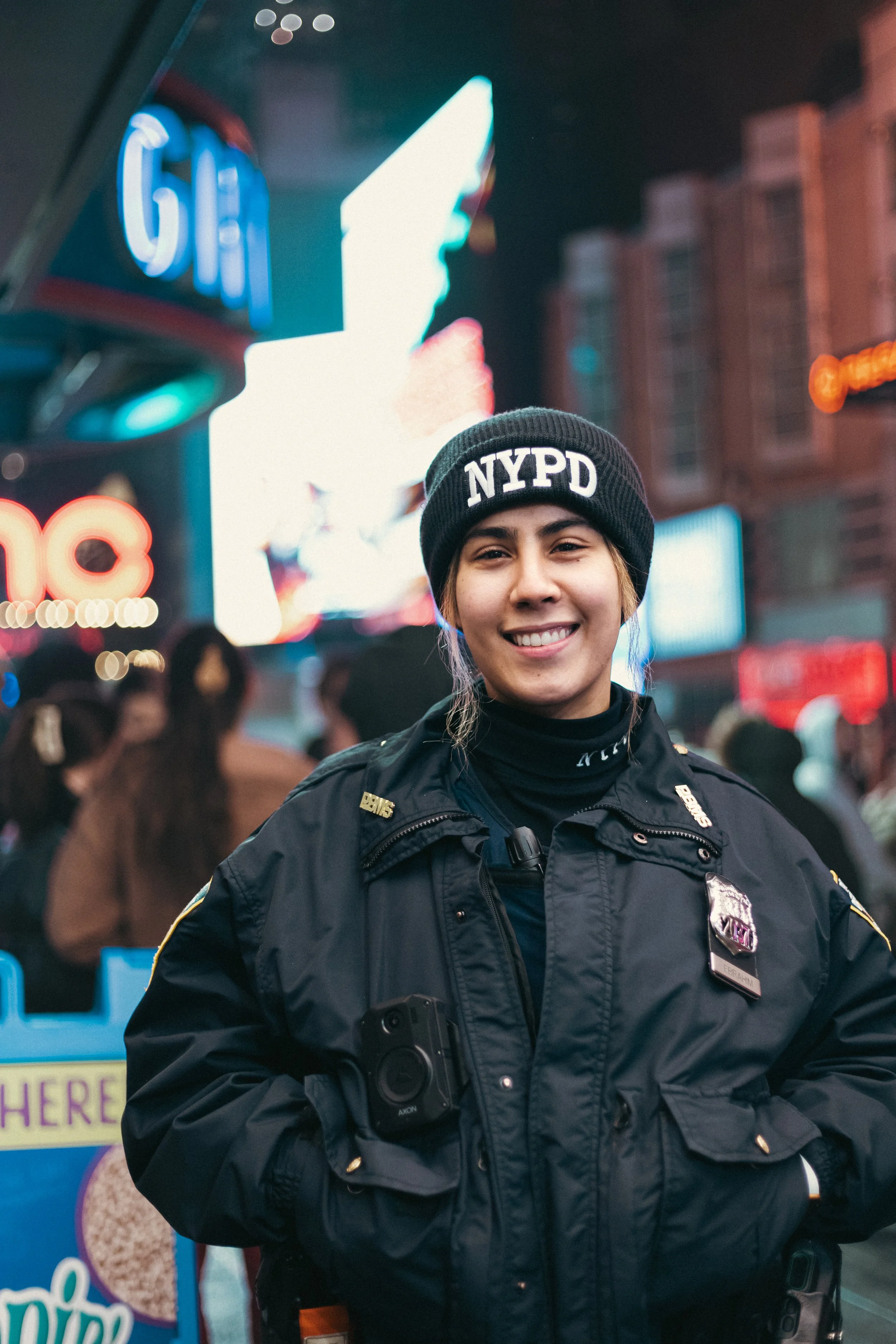 A smiling female NYPD officer in uniform standing in a busy city street at night, with neon signs and a crowd in the background.
