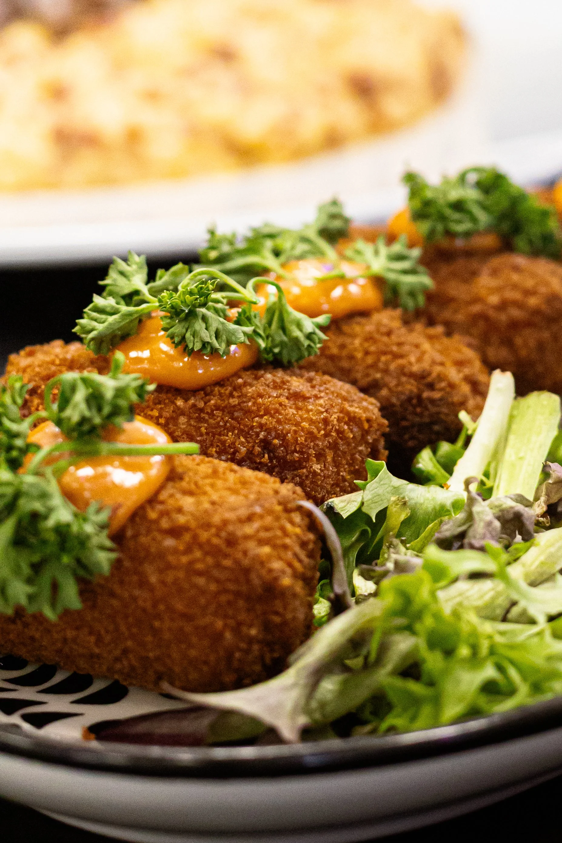 Close-up of fried food bites topped with a creamy orange sauce and garnished with cilantro, served with leafy greens on the side.