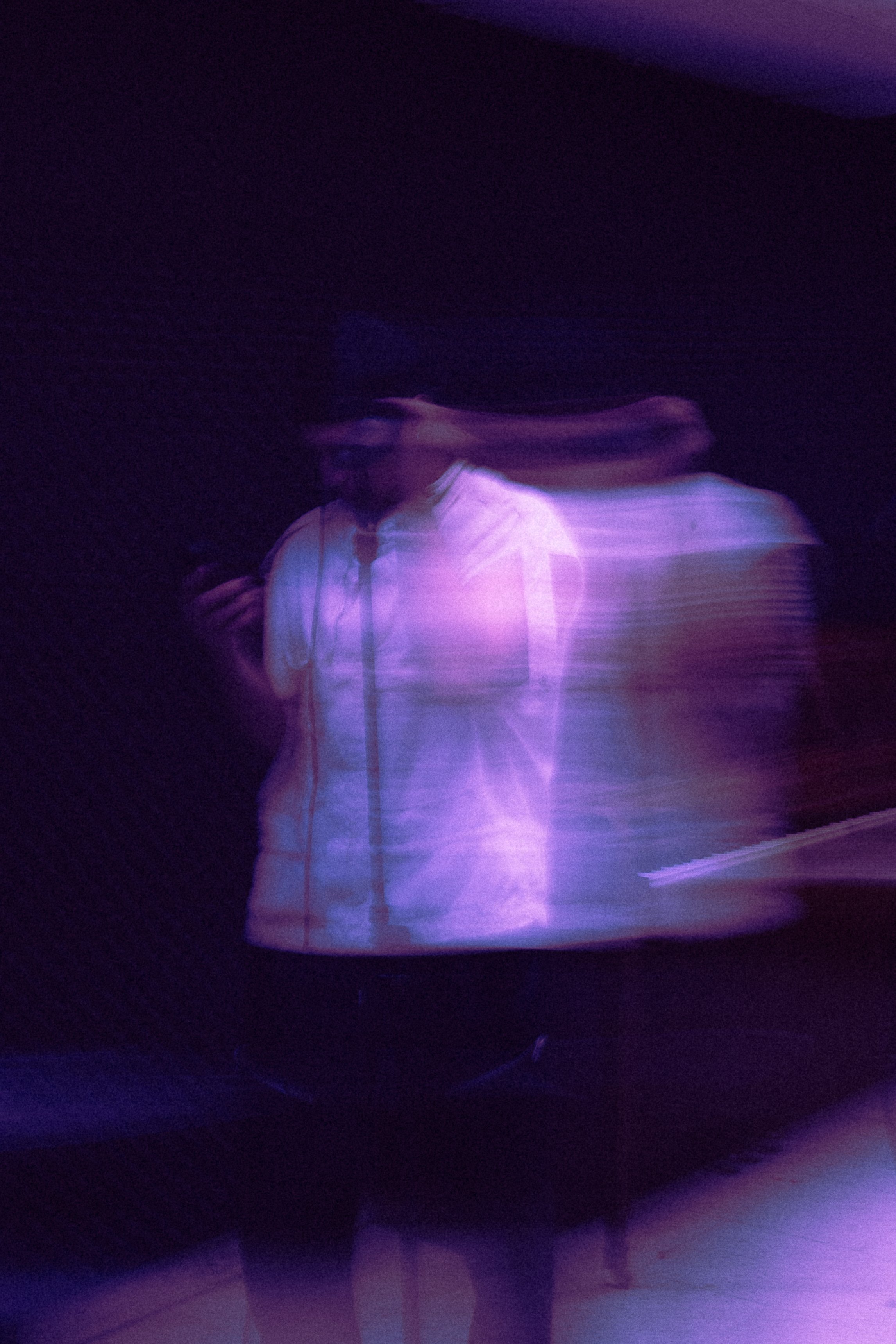 Blurry photo of two people in a dark room with purple lighting; one person is looking at their phone while the other is slightly behind.