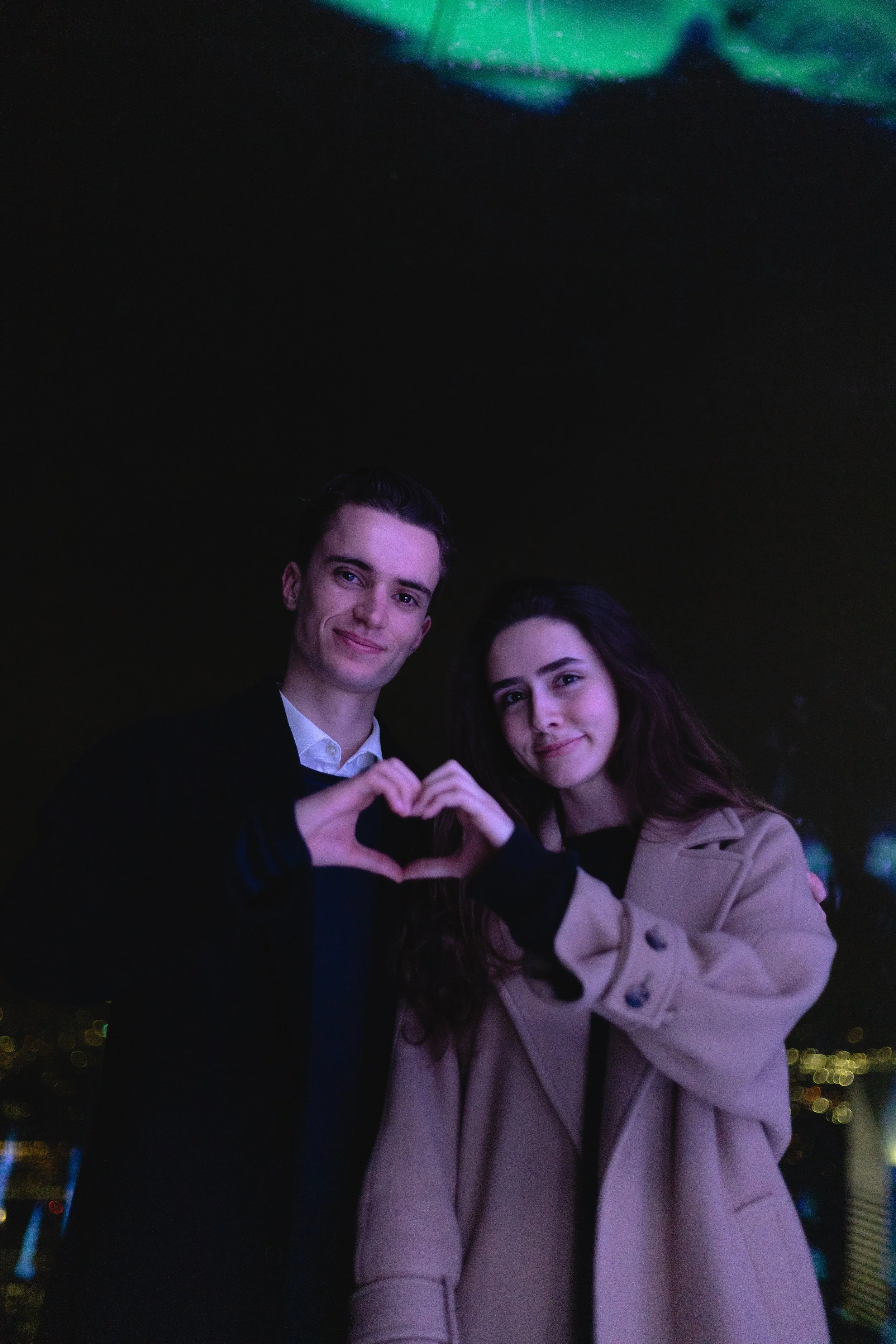 A young man and woman standing outdoors at night, forming a heart shape with their hands.