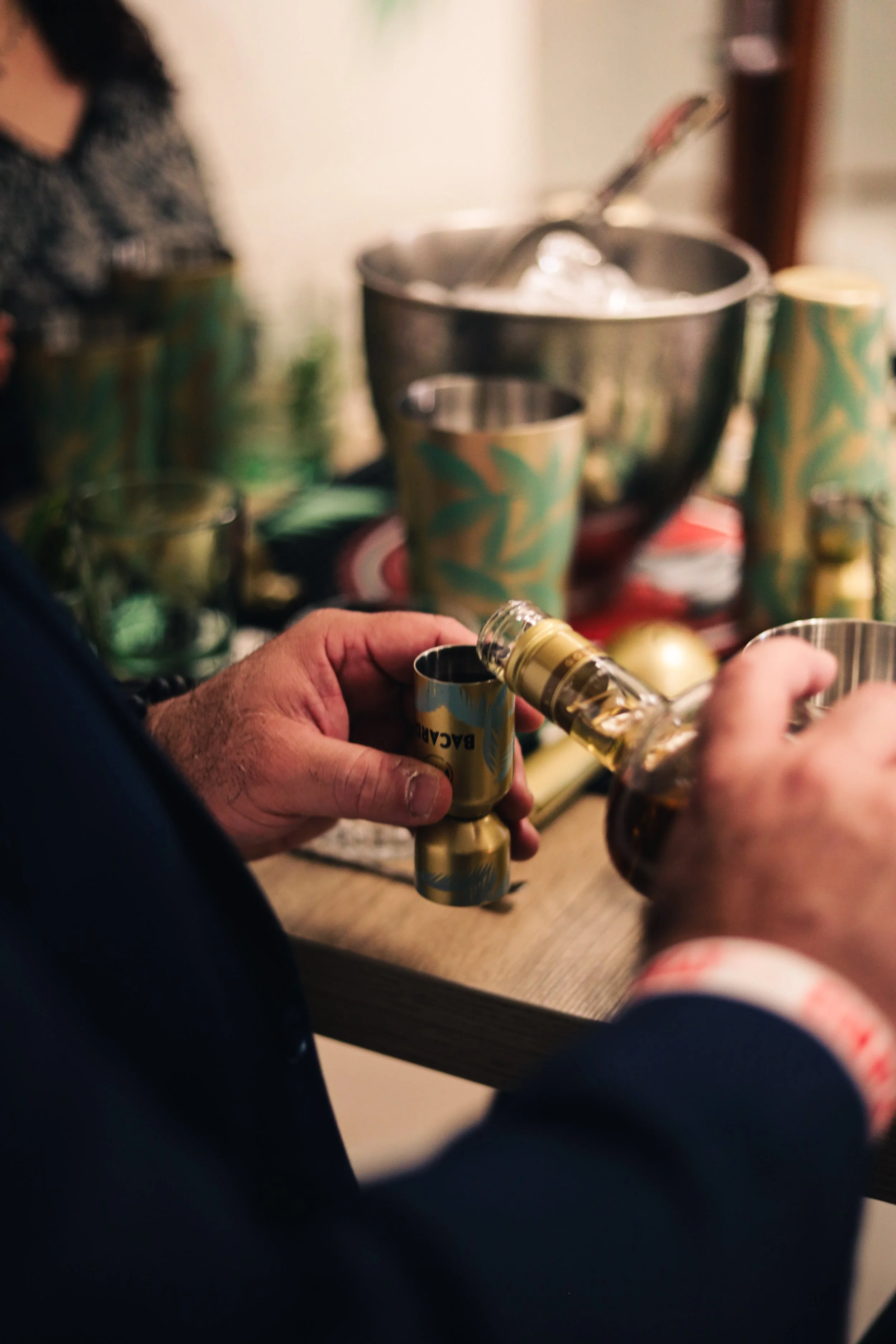 Close-up of a person pouring a dark liquor from a bottle into a small glass at a party. In the background, there are colorful cups and a metal bowl on a table.
