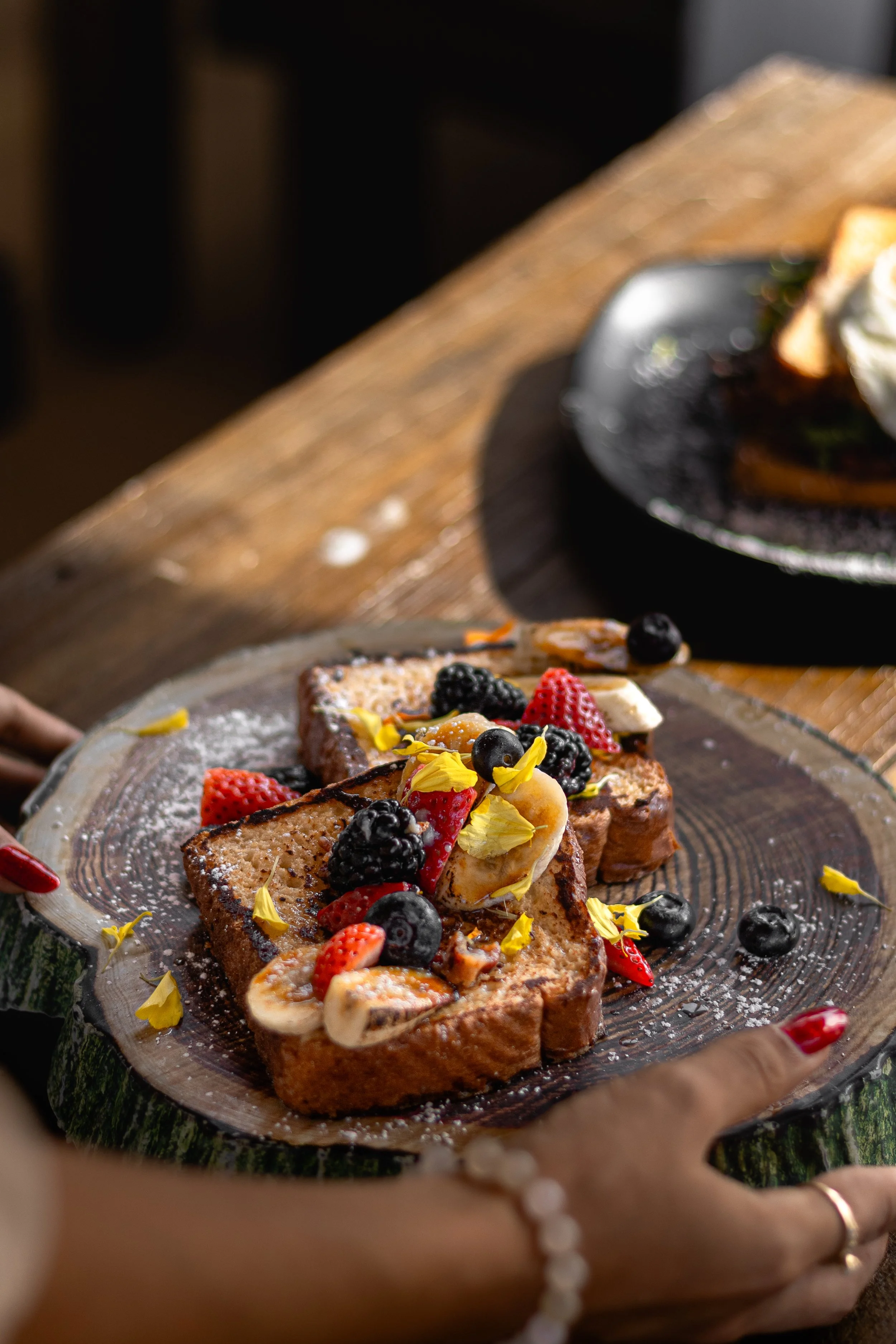 French toast topped with fresh berries, banana slices, edible flowers, powdered sugar, and syrup, served on a rustic wooden platter.