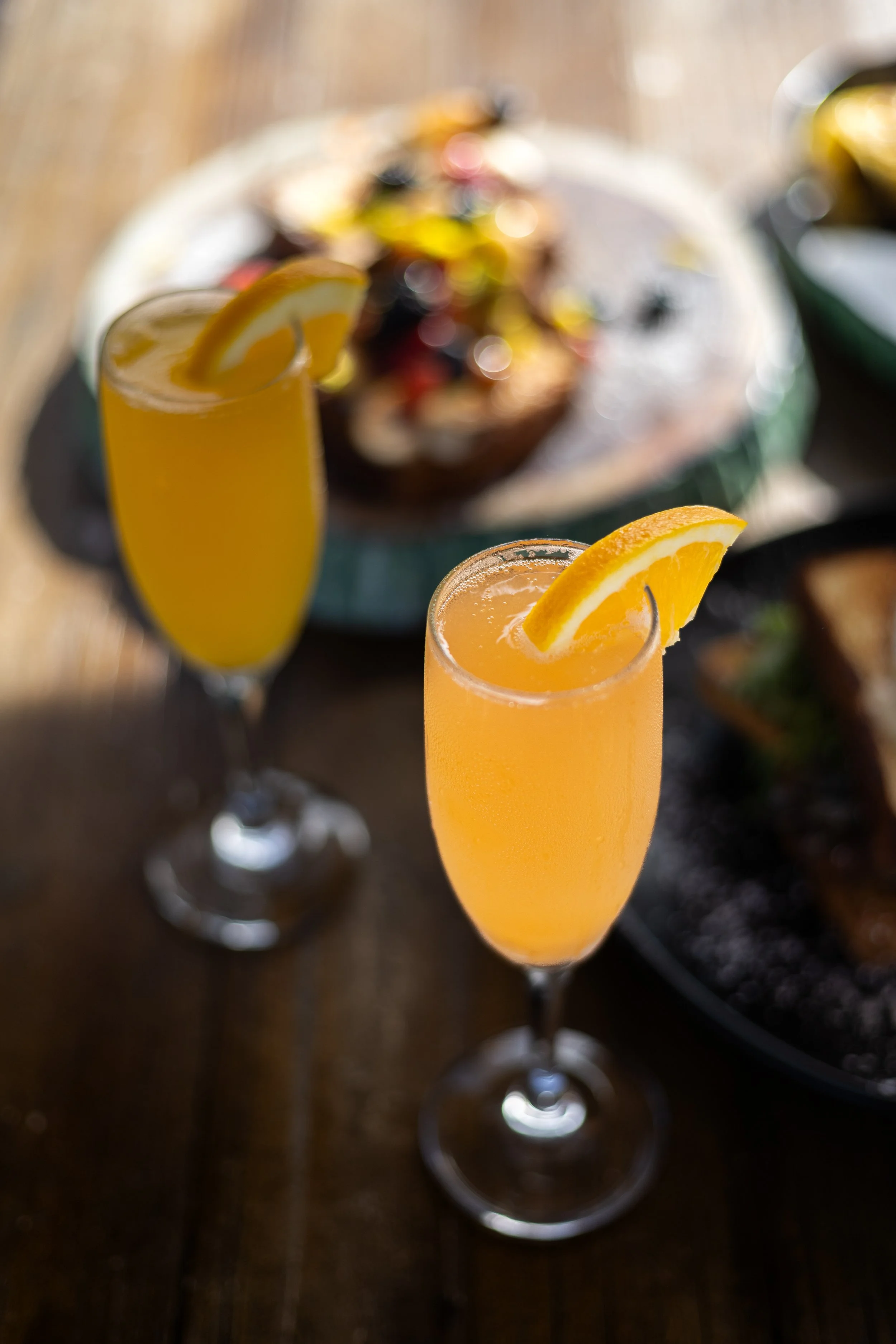 Two glasses of orange-colored beverages with lemon wedges on a dark wooden table, with blurred food dishes in the background.