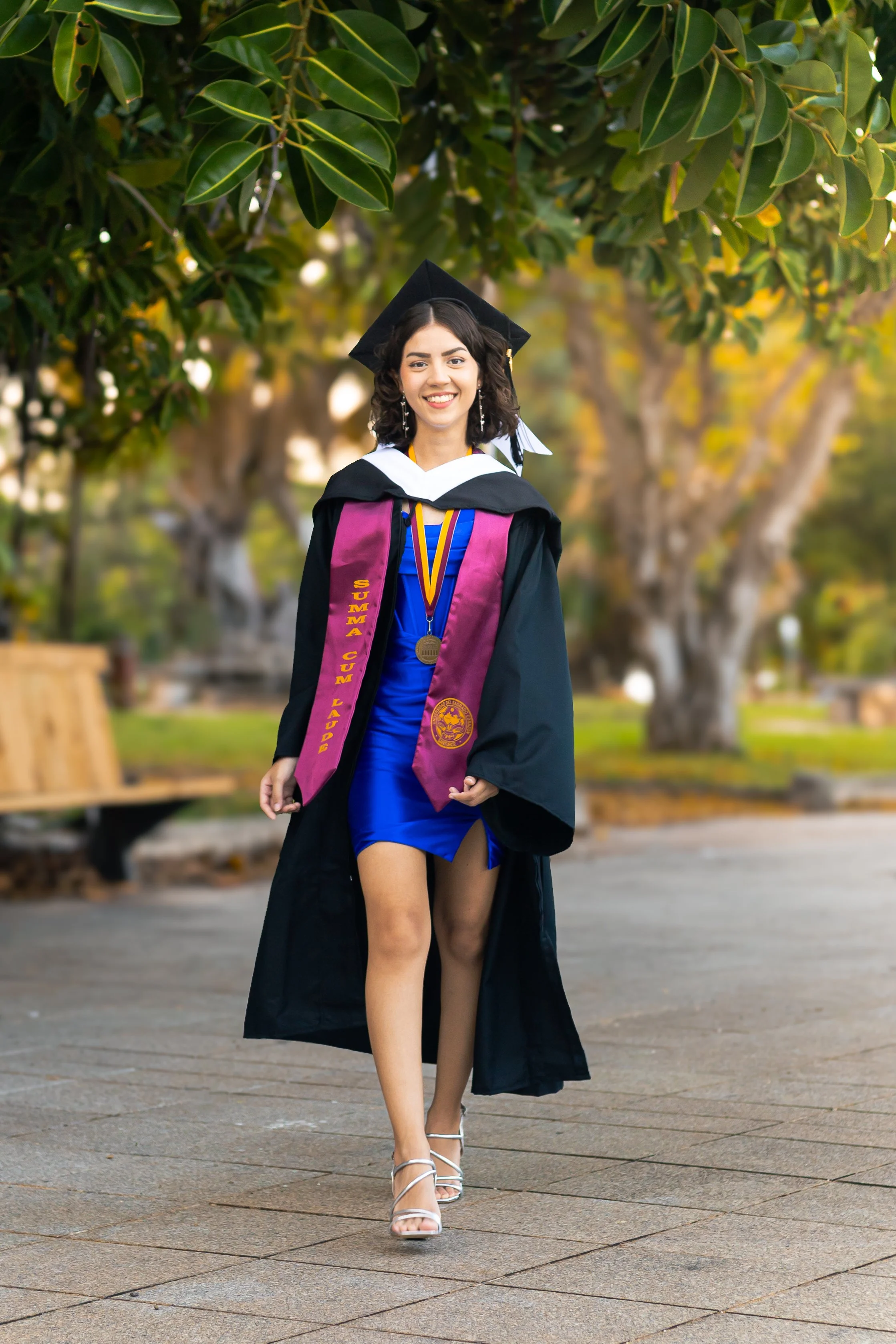 A young woman in a graduation cap and gown, smiling, walking outdoors on a paved path with trees and benches in the background.