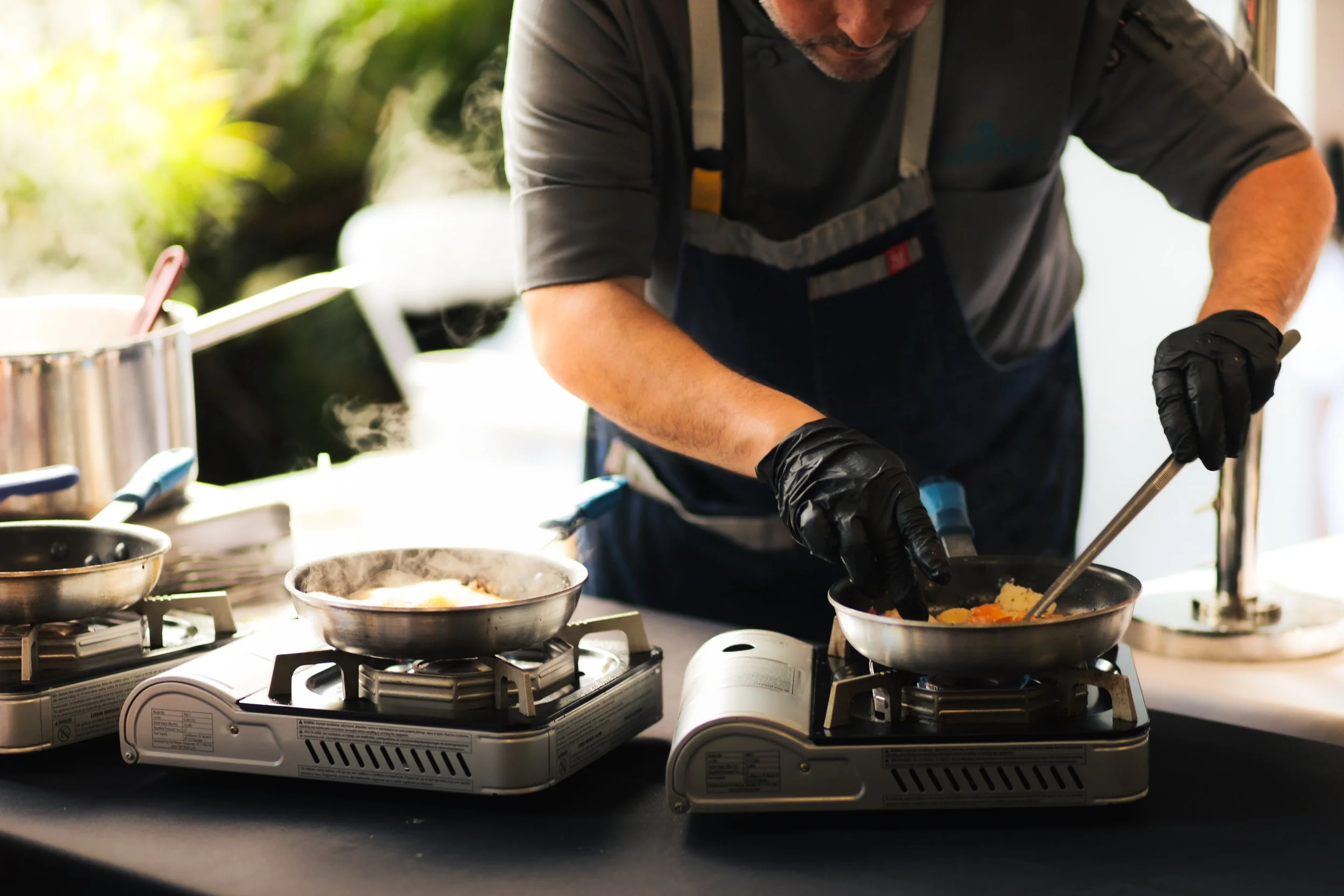 A man cooking food on two portable gas stoves, using tongs to stir ingredients in a frying pan. He is wearing black gloves, a gray shirt, and a navy apron. Cooking utensils and ingredients are on the black table, with steam rising from the pans.