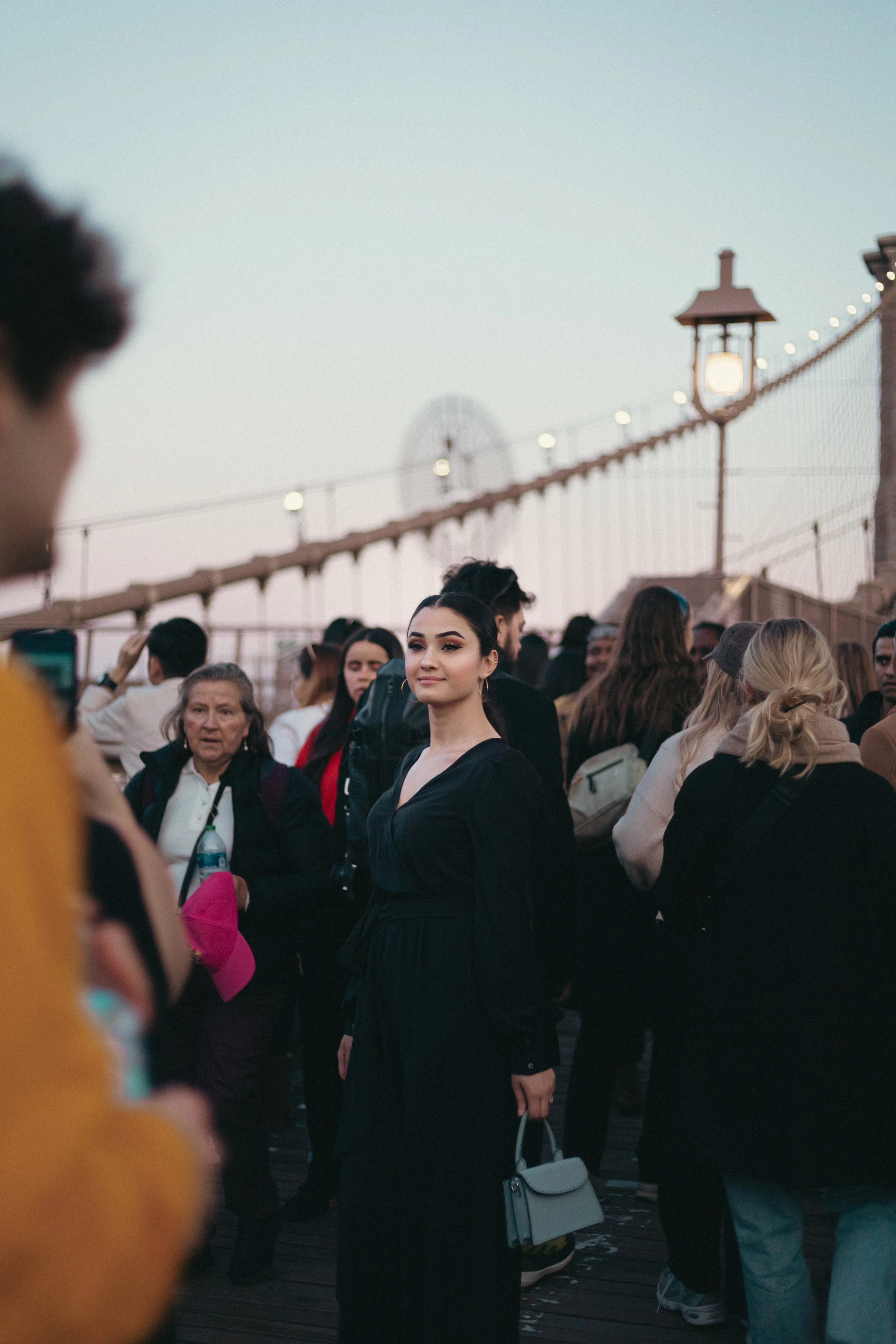 A young woman in a black dress holding a small white purse stands on a bridge with a crowd of people around her, a Ferris wheel visible in the background at sunset.