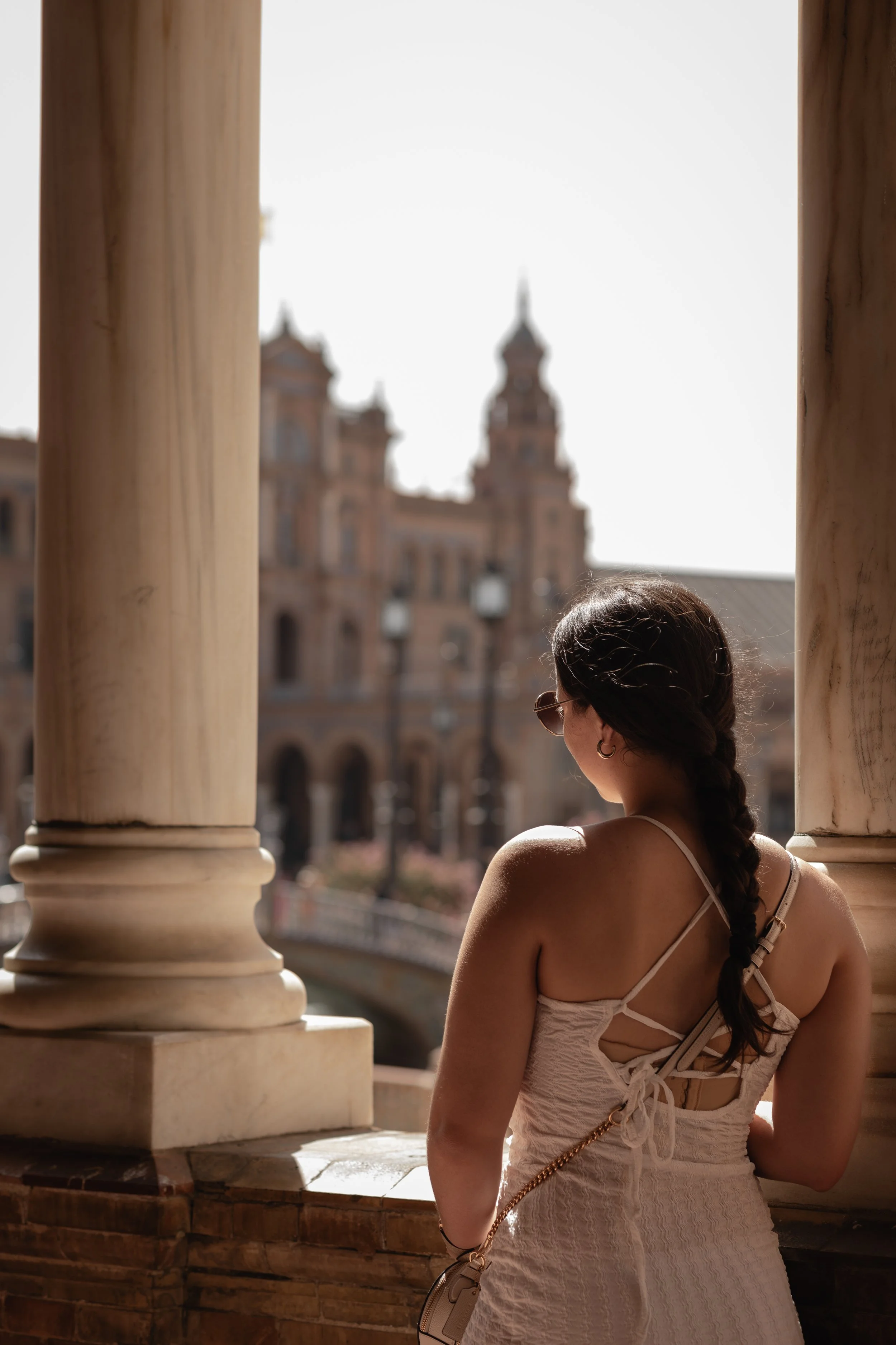 A woman with dark hair in a braid, wearing sunglasses and a beige dress, stands on a balcony with large stone columns, looking at a historic building with towers in the background.