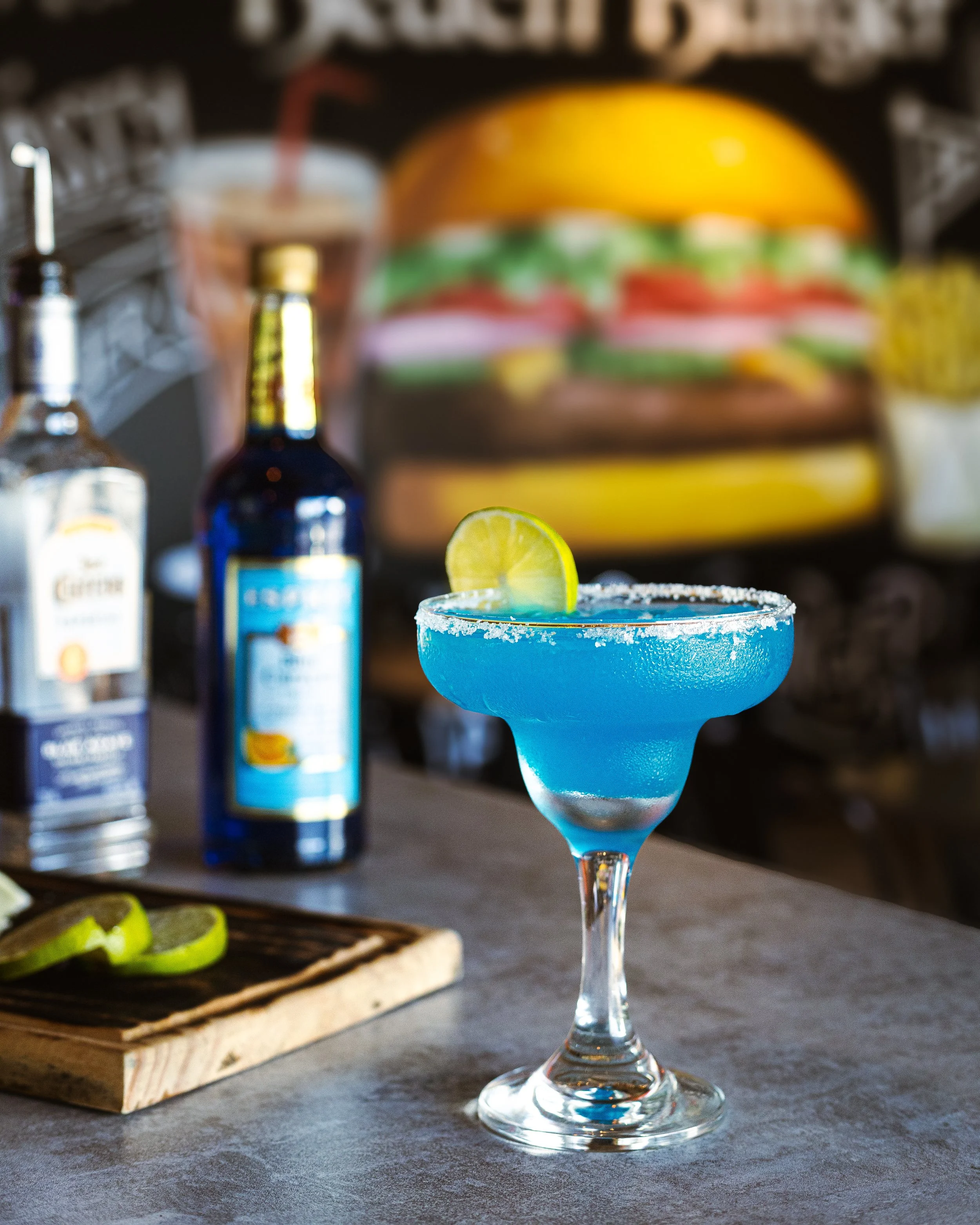 A blue margarita with a salted rim and a lemon slice garnish on a bar counter, with bottles of alcohol and a large hamburger illustration in the blurred background.