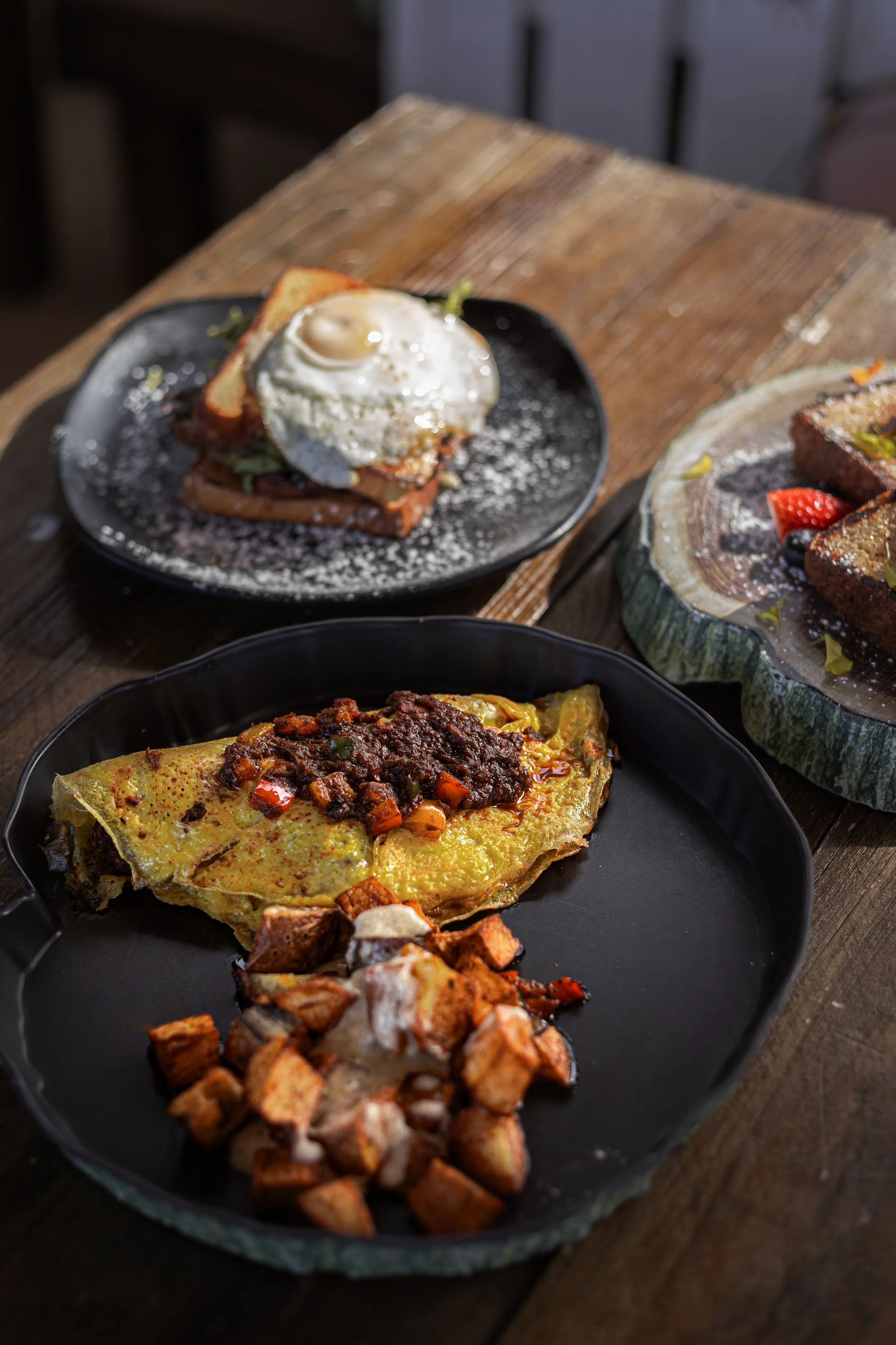 A rustic wooden table with three plates of breakfast food: an omelet with sausage and diced vegetables, topped with a meat sauce; a plate with toast, fried egg, and leafy greens; and a slice of cake with strawberries.