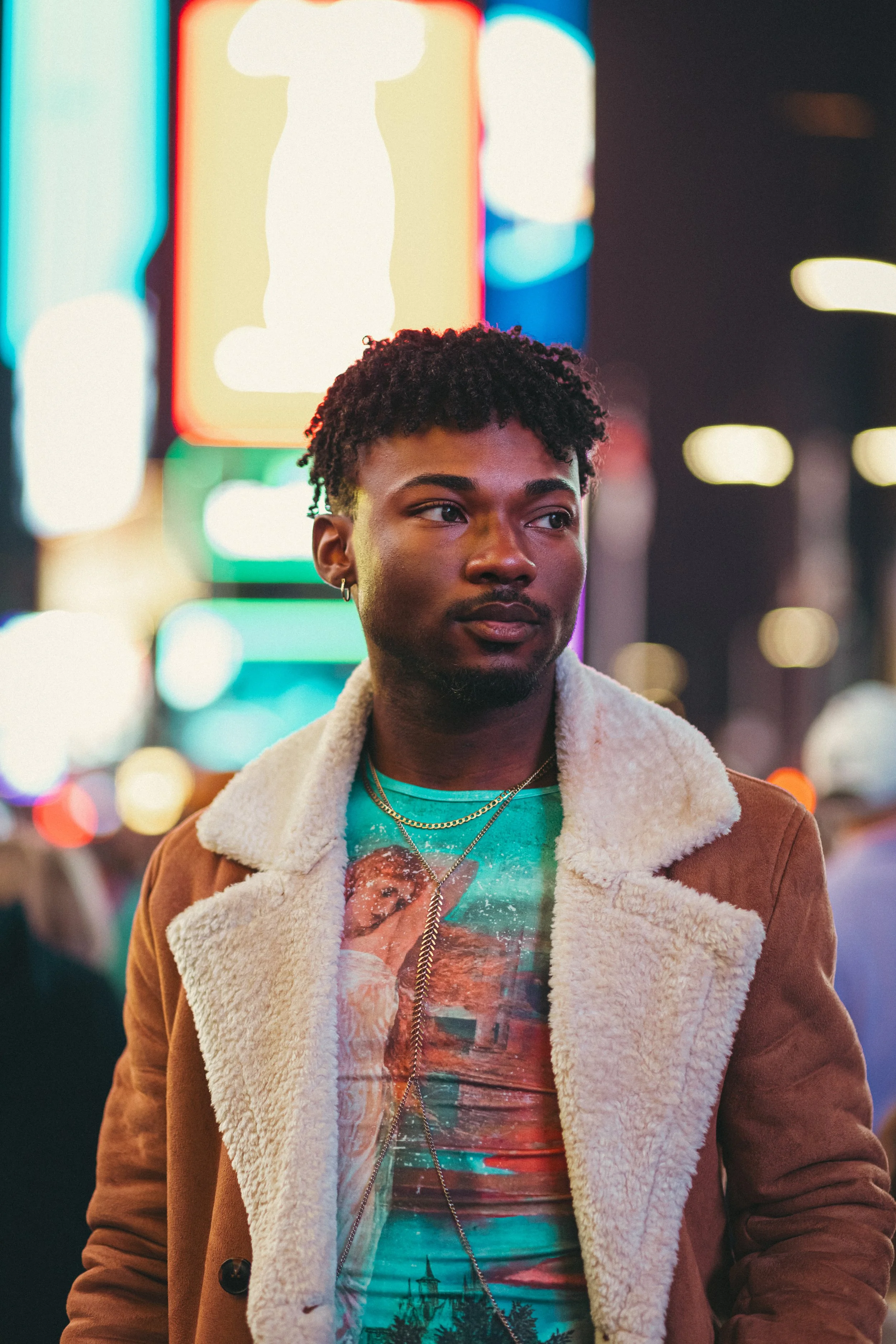 A young man with curly hair and a beard, wearing a brown jacket with a shearling collar over a colorful graphic t-shirt, stands on a city street at night illuminated by neon signs and bright city lights.