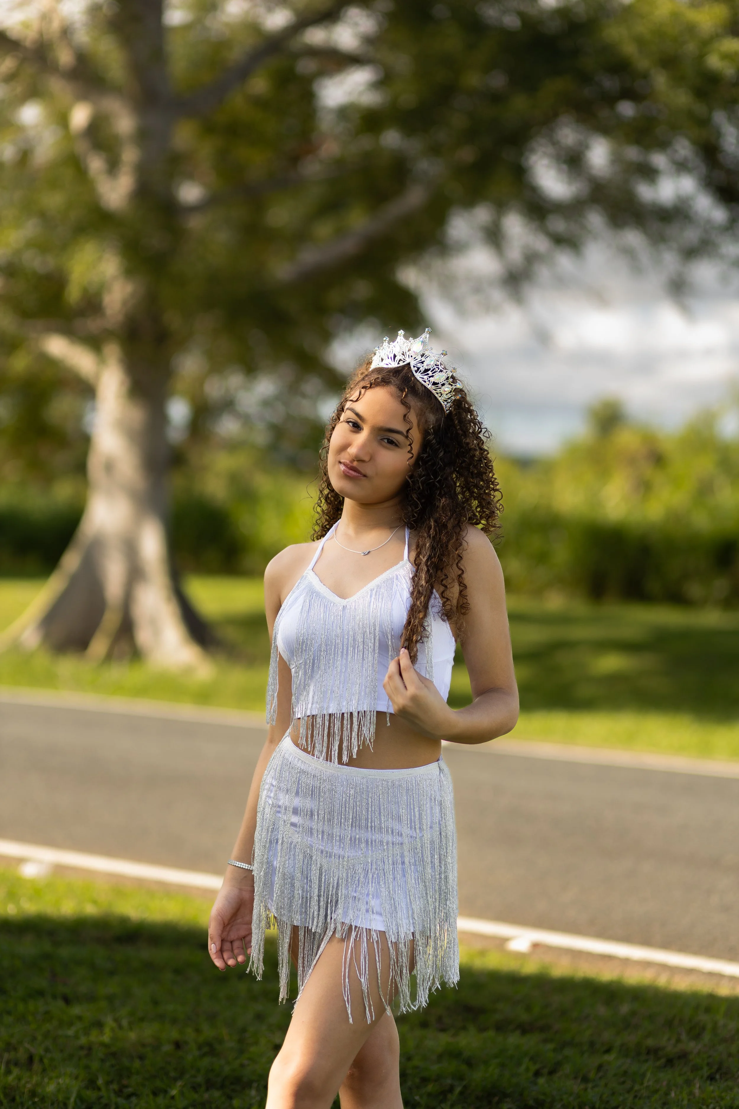 Young woman wearing a silver tiara and matching fringe dress standing outdoors near a tree.