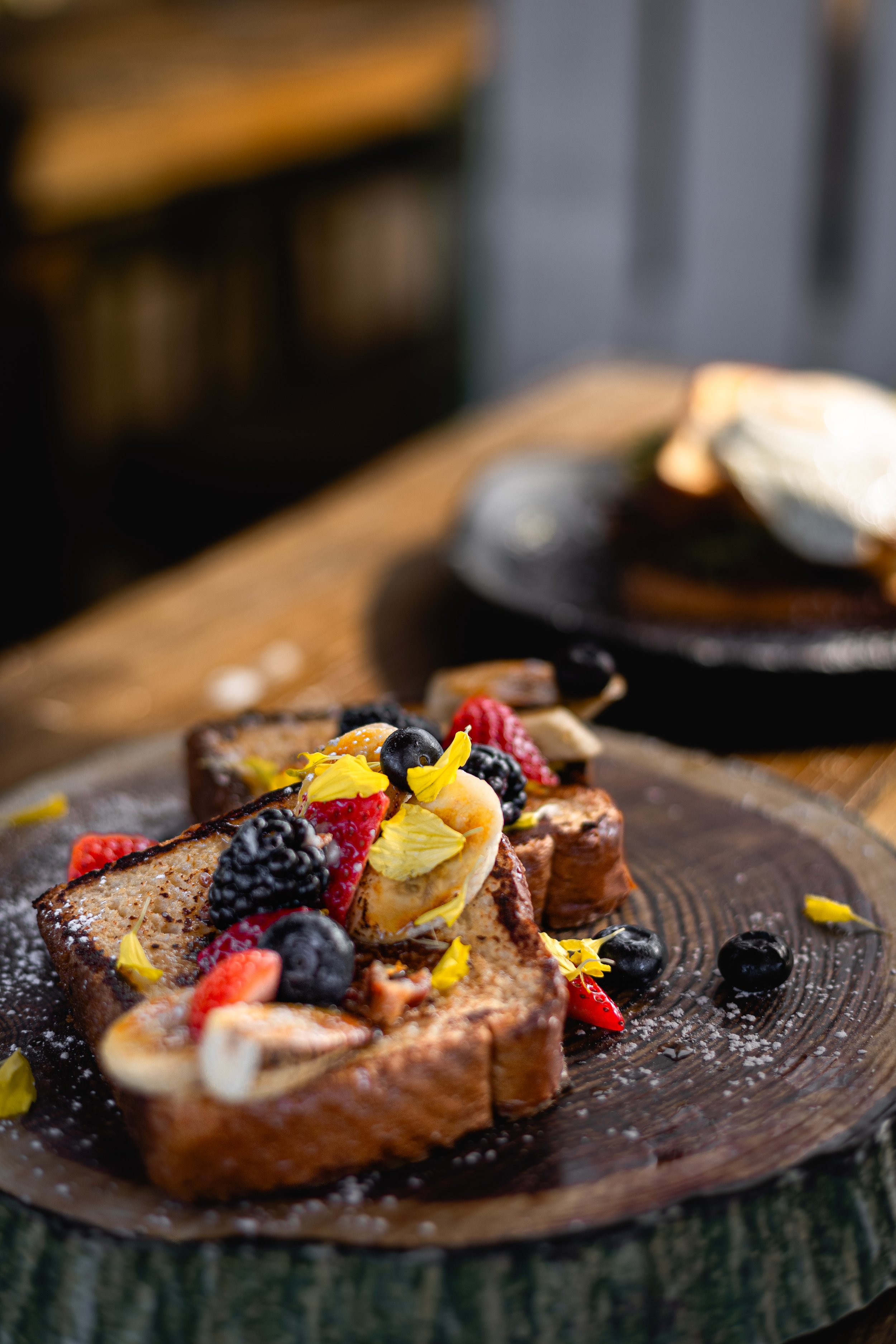 French toast topped with berries, banana slices, edible flower petals, and powdered sugar on a wooden platter.