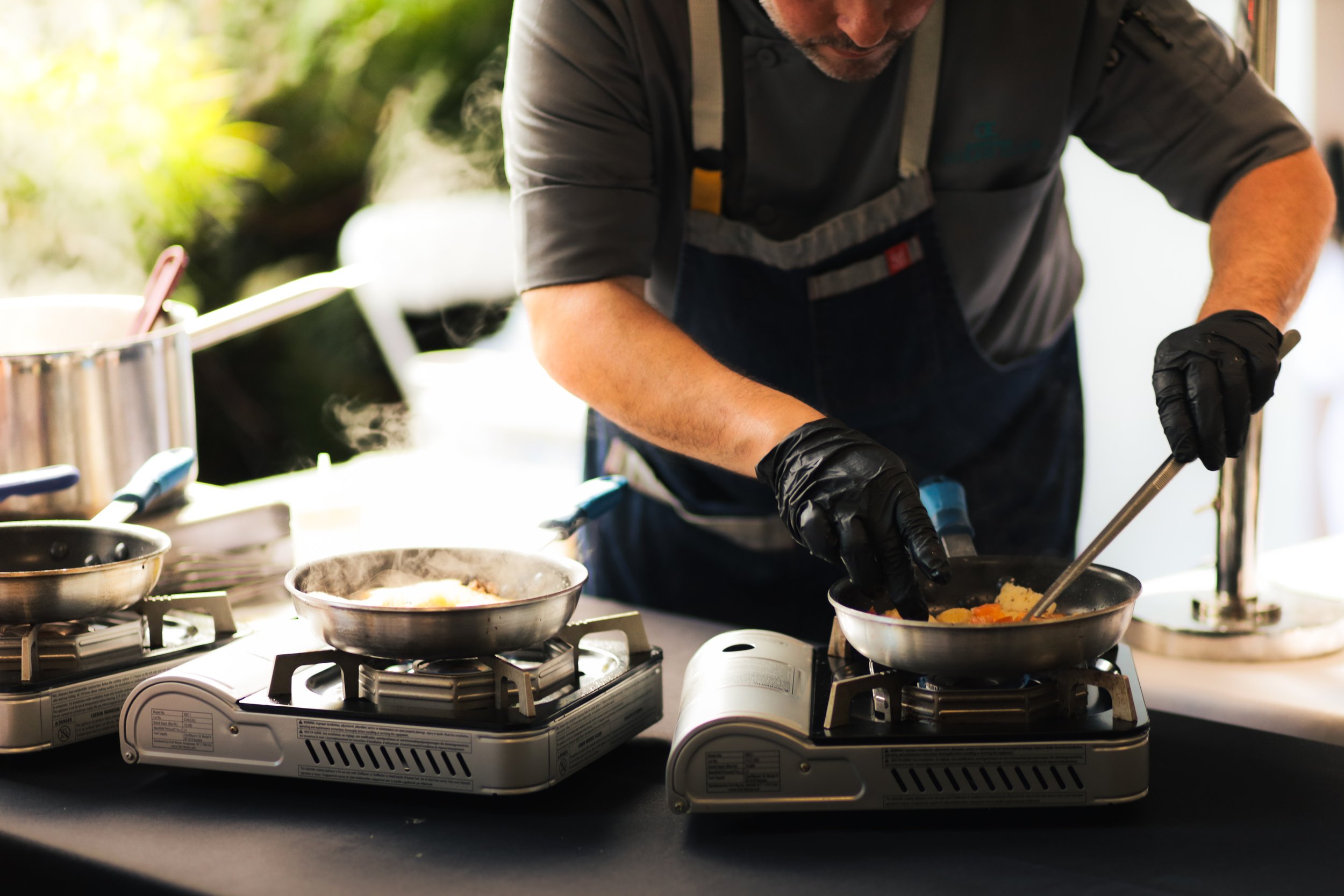 A man wearing black gloves preparing food on portable gas stoves during outdoor cooking, with steam rising from the pans.