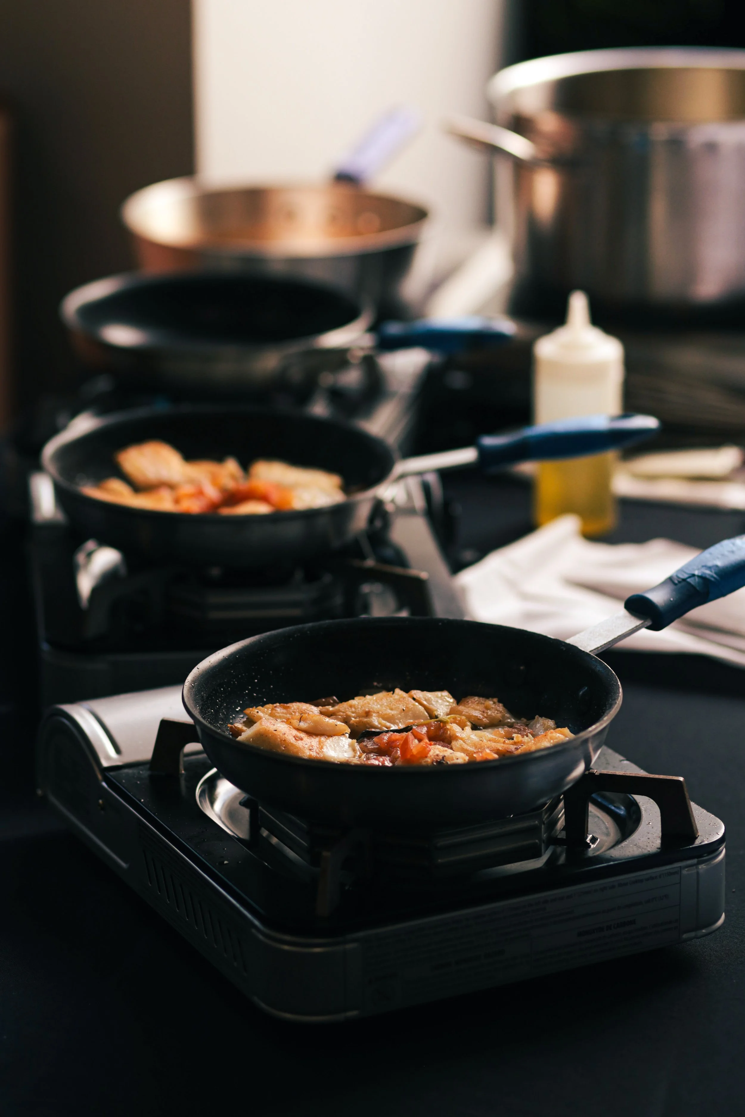 Multiple frying pans cooking food on a portable gas stove, with ingredients and utensils nearby in a kitchen setting.