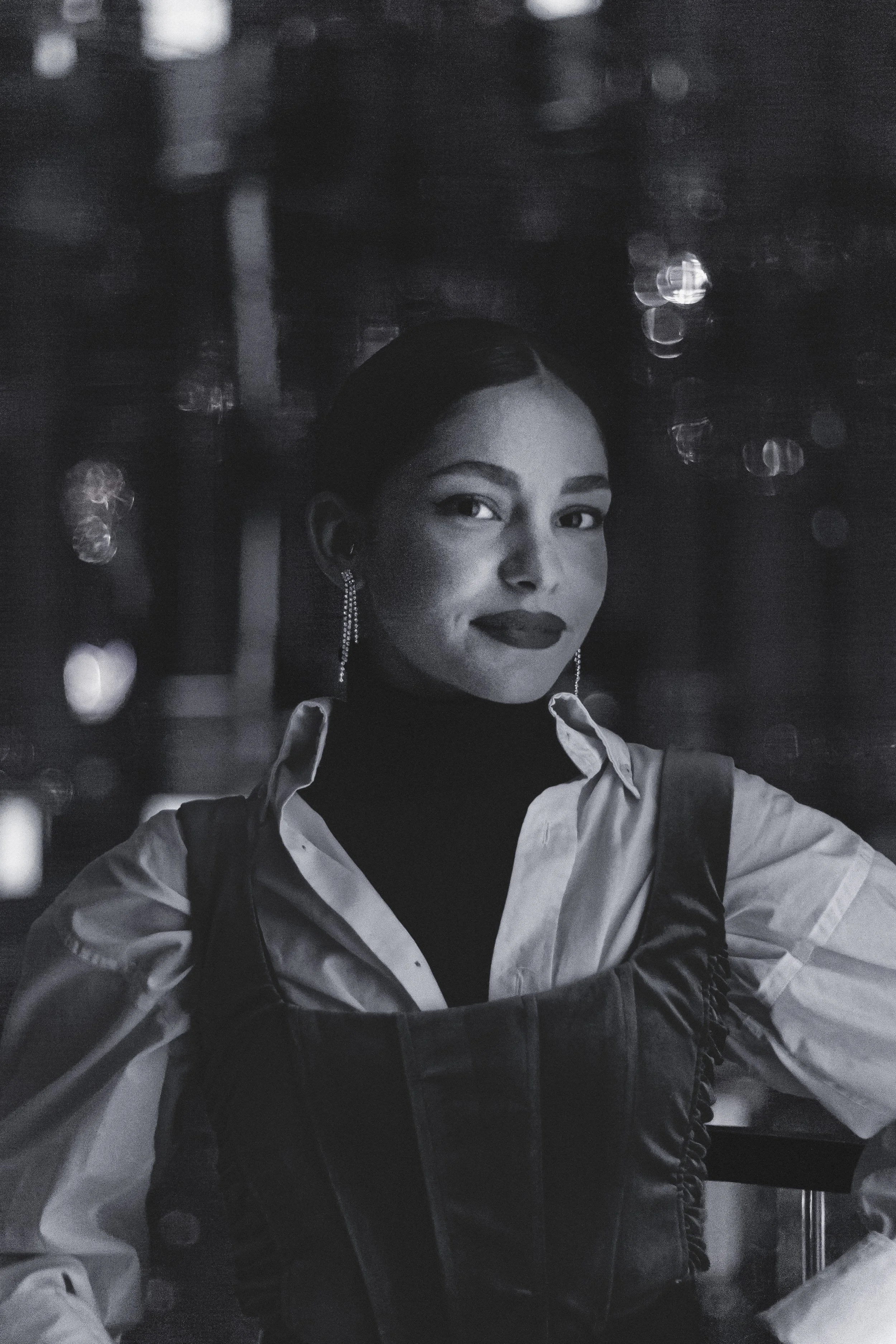 Portrait of a woman in black and white with earrings, wearing a layered outfit, in a dimly lit environment with bokeh effects in the background.