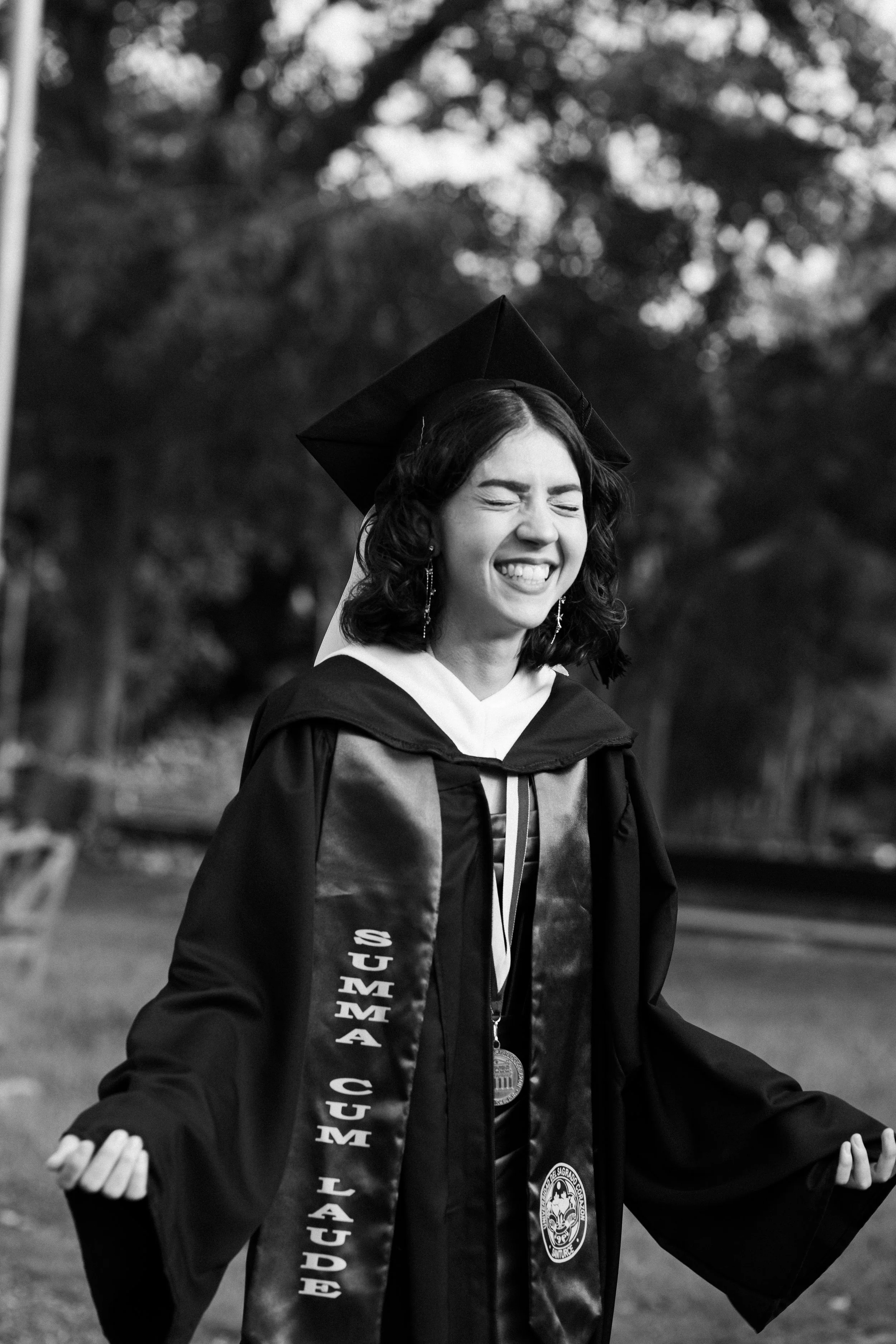 Black and white photo of a smiling young woman in graduation gown and cap, with her eyes closed, outdoors with trees in the background.