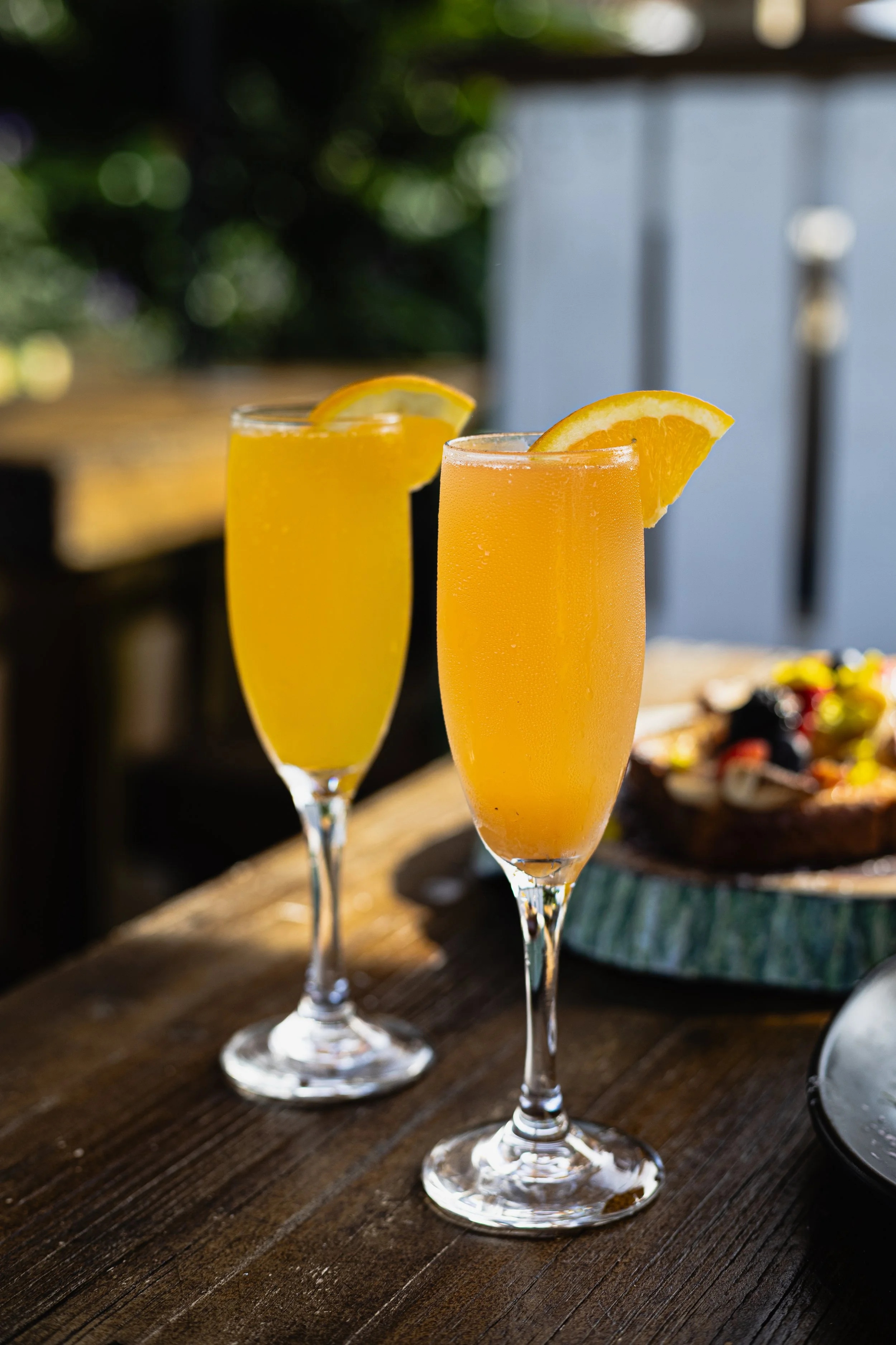 Two champagne flutes with orange juice garnished with orange slices on a wooden table outside, with a platter of food in the background.
