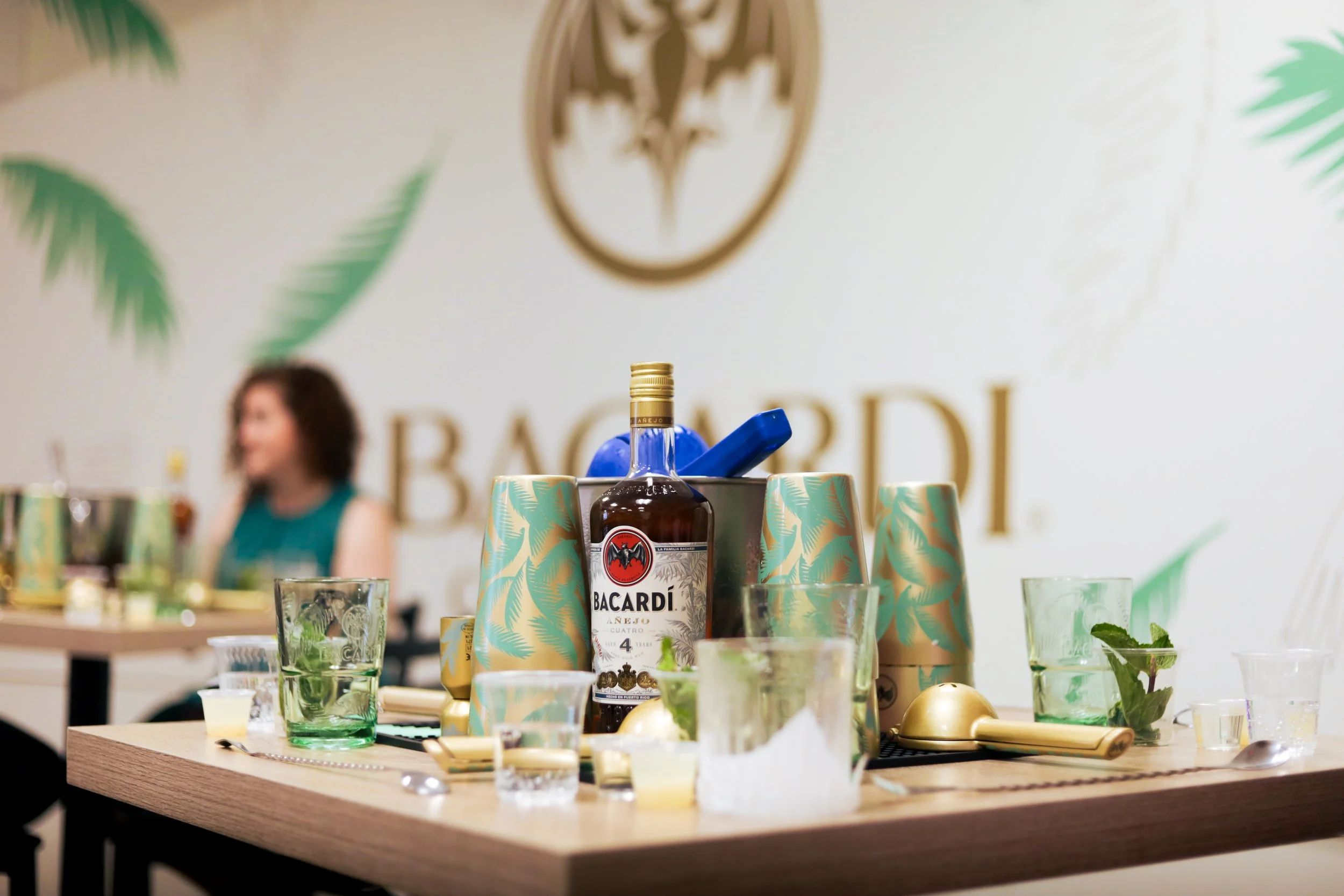Bar setup with Bacardi rum bottle, paper cups, glasses with mint leaves, and cocktail utensils on a wooden table in front of a wall with a Bacardi logo and decorative palm leaves.