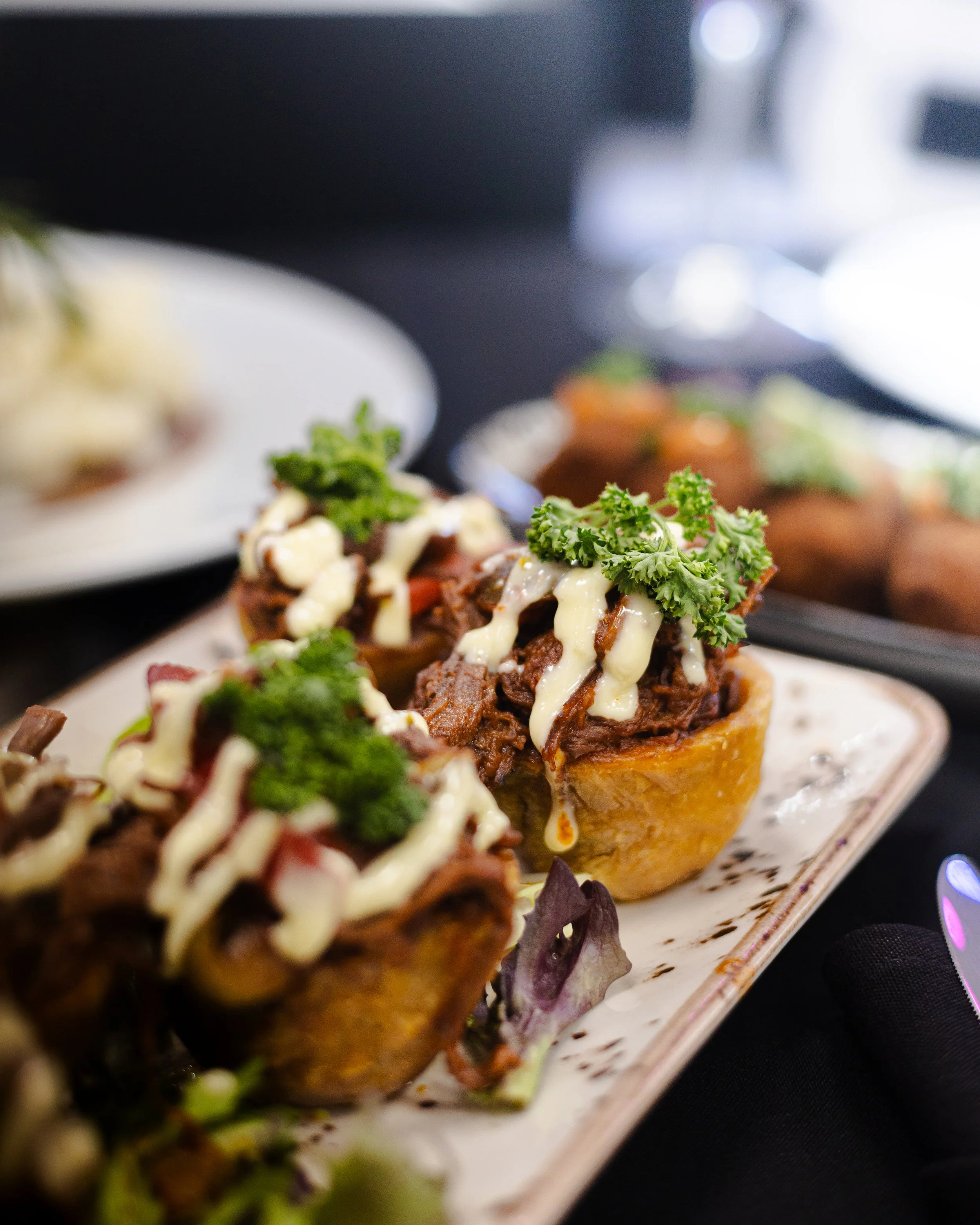 Close-up of three bun cupcakes topped with shredded meat, white drizzle, and parsley on a rectangular plate, with a blurred background of additional food.
