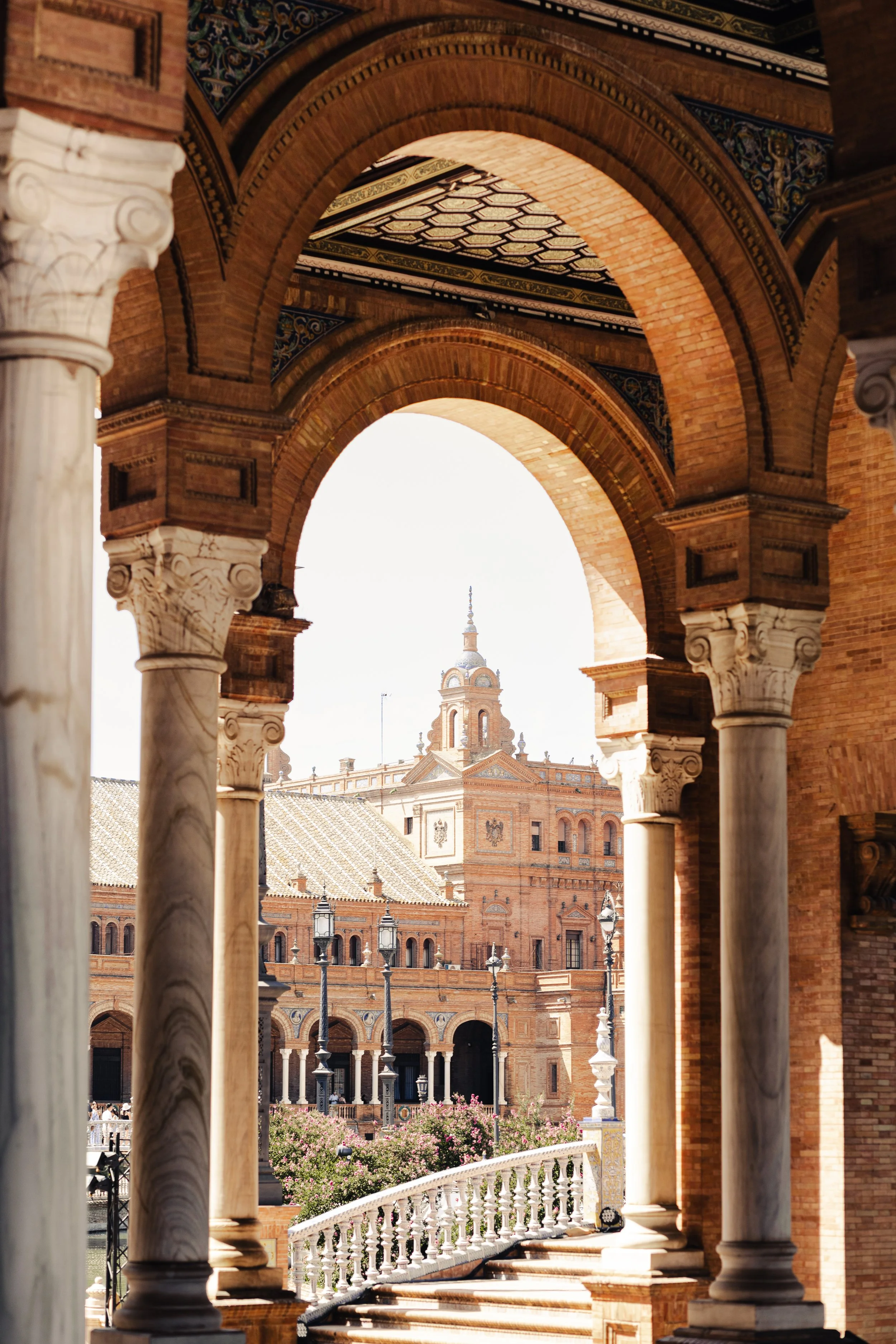 View of a historic brick building with a dome and tower, seen through stone columns and arches of a classical-style structure, with a decorative railing and flowering bushes in the foreground.