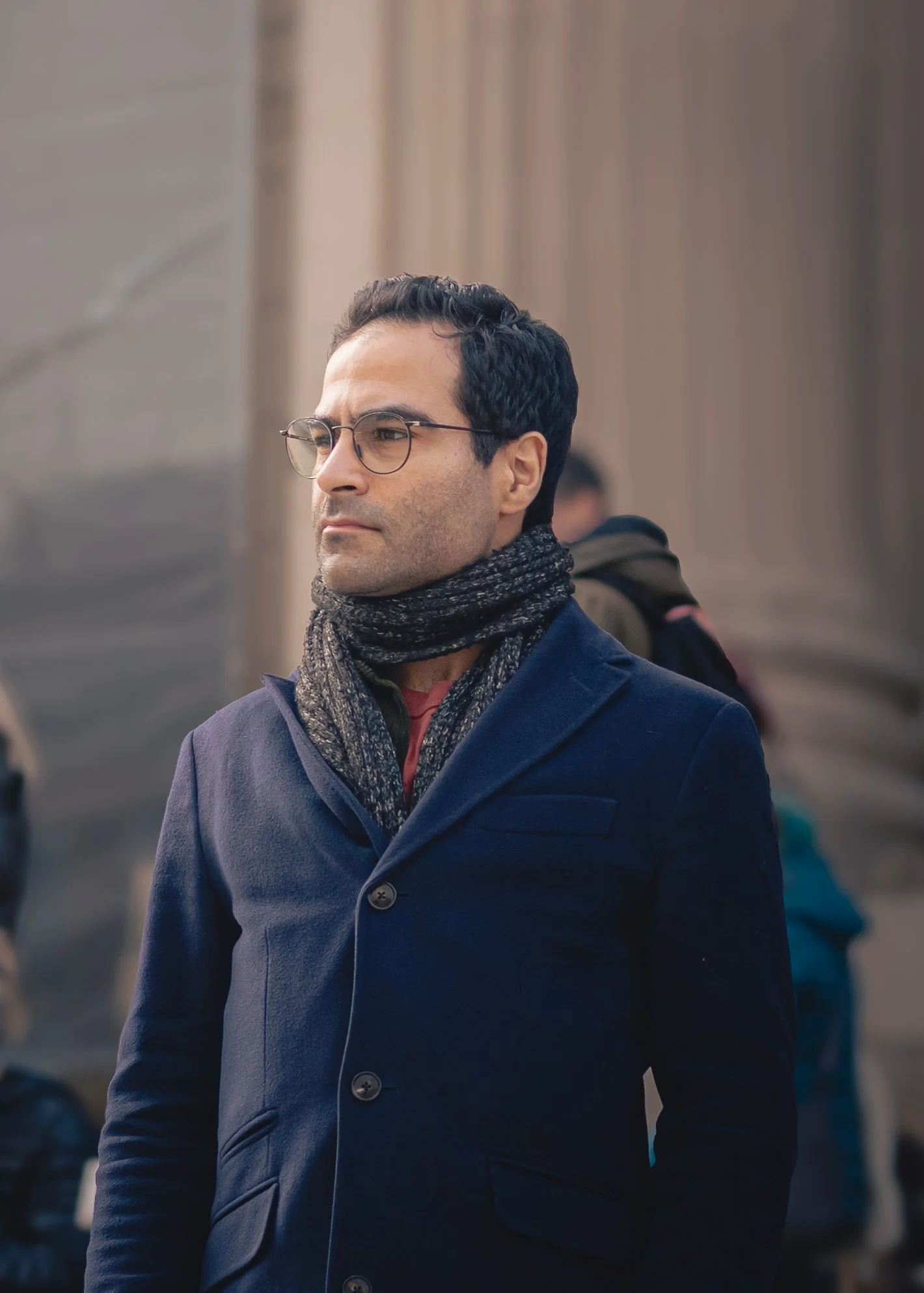 A man wearing glasses, a dark blue coat, and a patterned scarf standing outdoors in front of a stone building, with a serious expression.