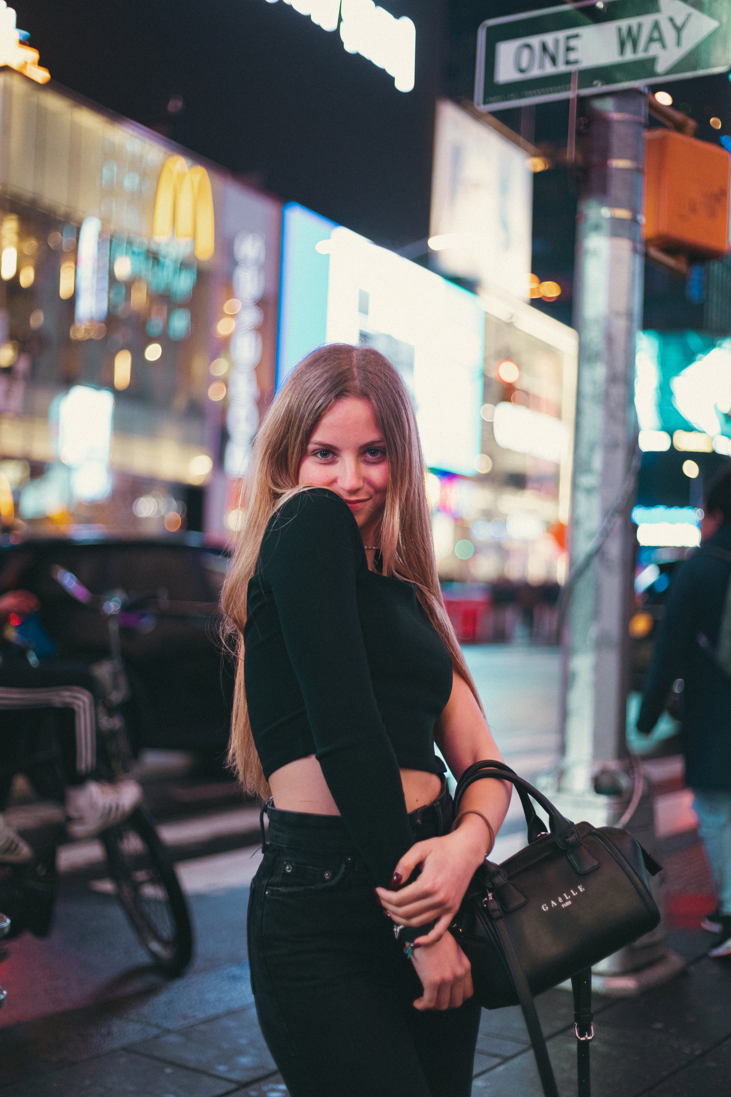 A young woman standing on a city street at night with bright lights and billboards in the background, holding a black handbag.