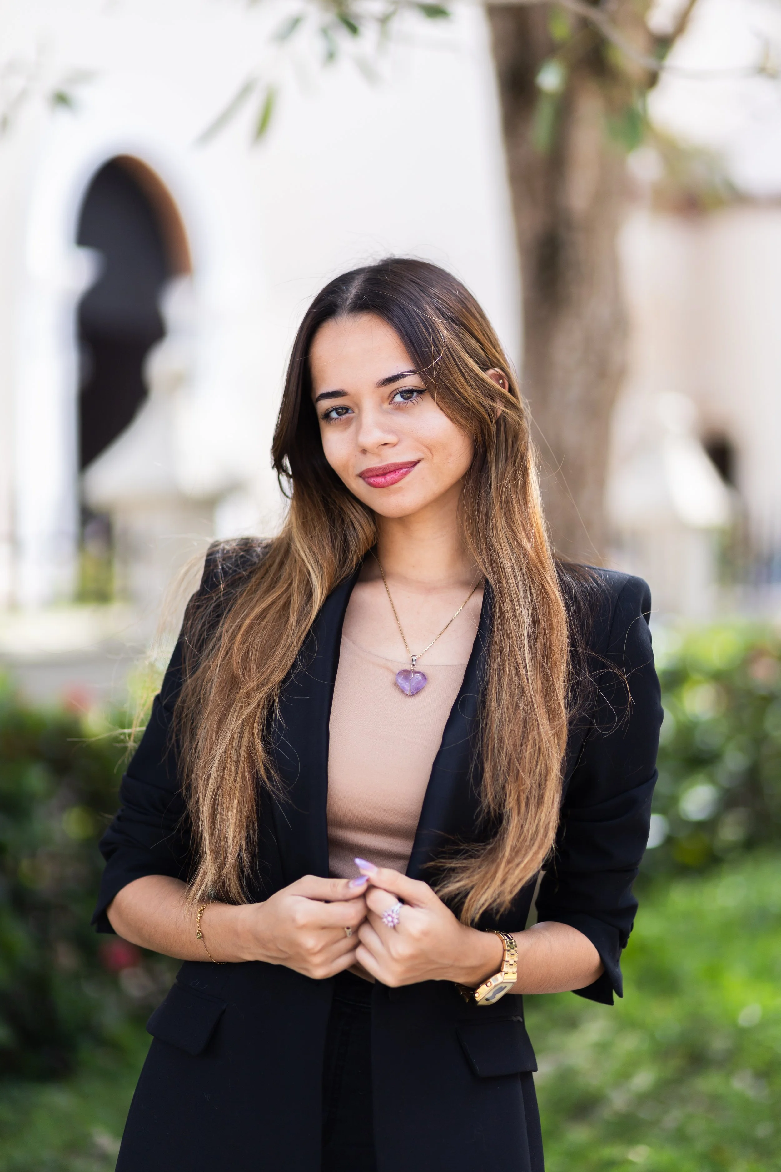 A young woman with long, wavy brown hair and light skin standing outdoors, wearing a black blazer, beige top, and jewelry including a purple heart necklace and gold watch, with a blurred background of greenery and a white building.