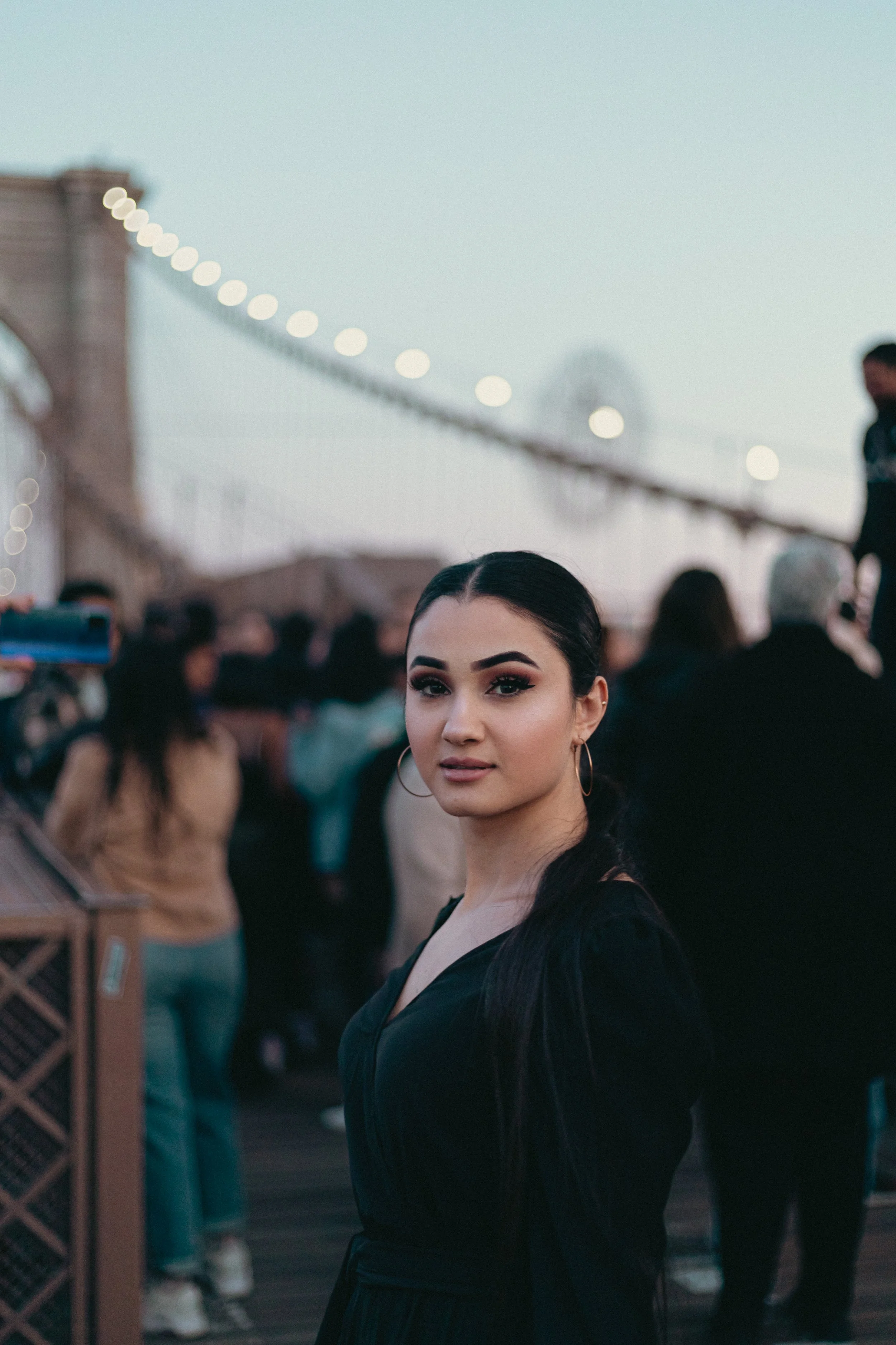 A young woman with dark hair and hoop earrings looking at the camera, at an outdoor event or fair during the evening, with a blurred crowd in the background and a bridge with lights.