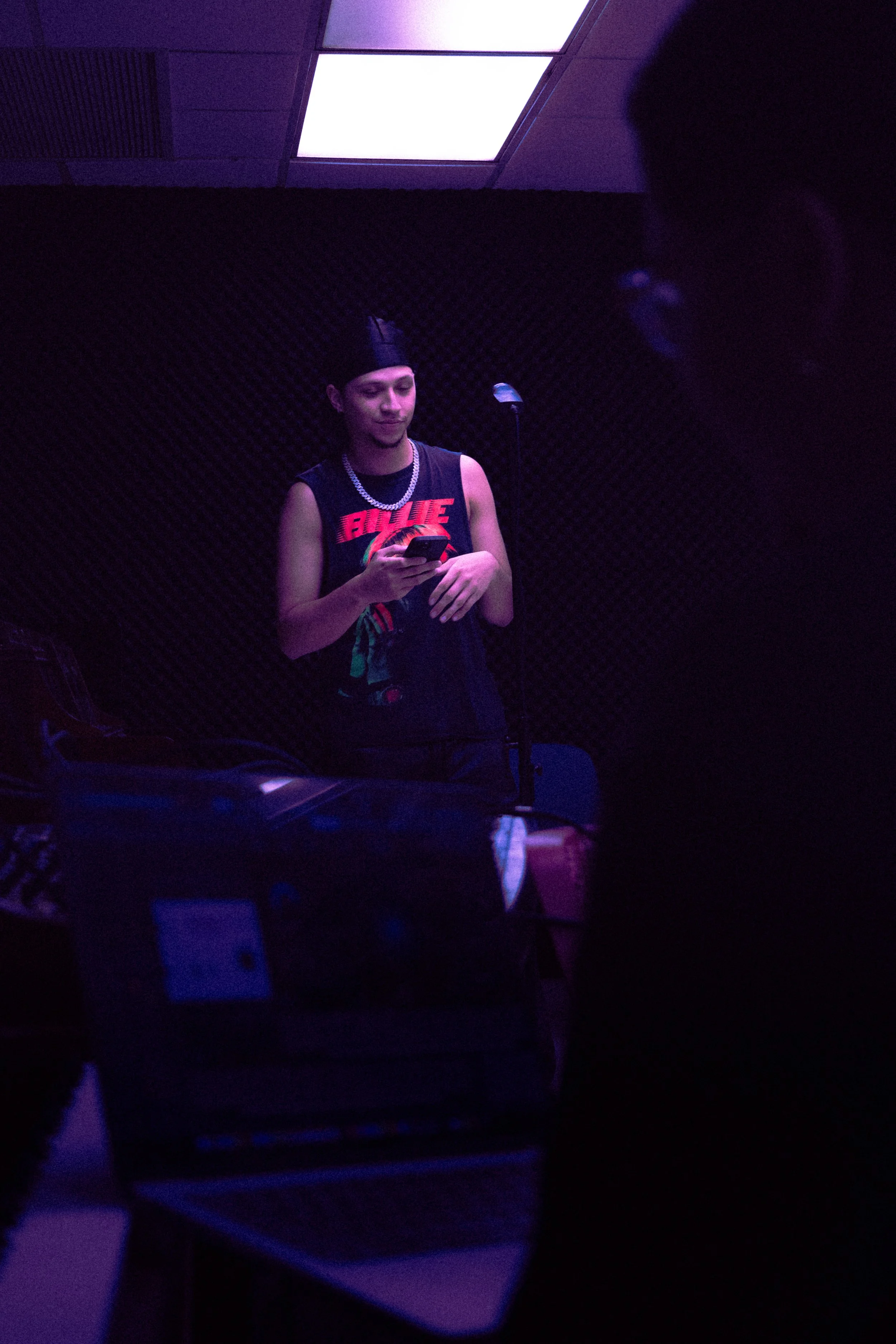 A young man with a bandana and jewelry standing in a dimly lit room, looking at his phone, with a microphone and equipment nearby, illuminated by purple lighting.