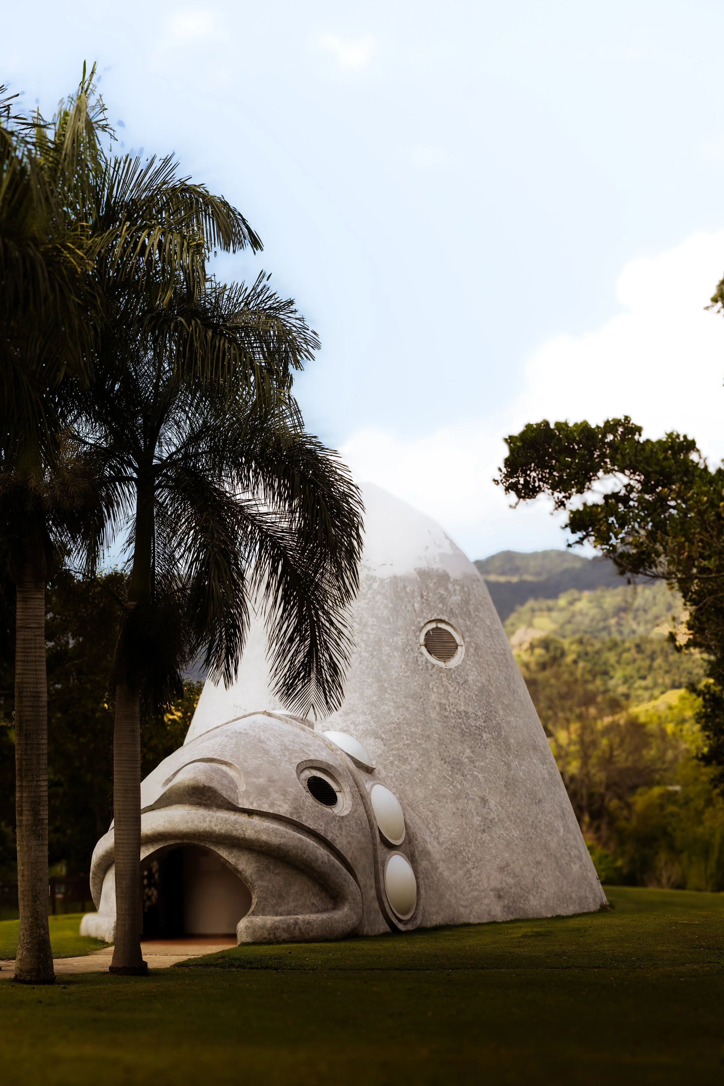 Unique building resembling a fish with an open mouth, circular windows, surrounded by palm trees and greenery, mountains in the background.