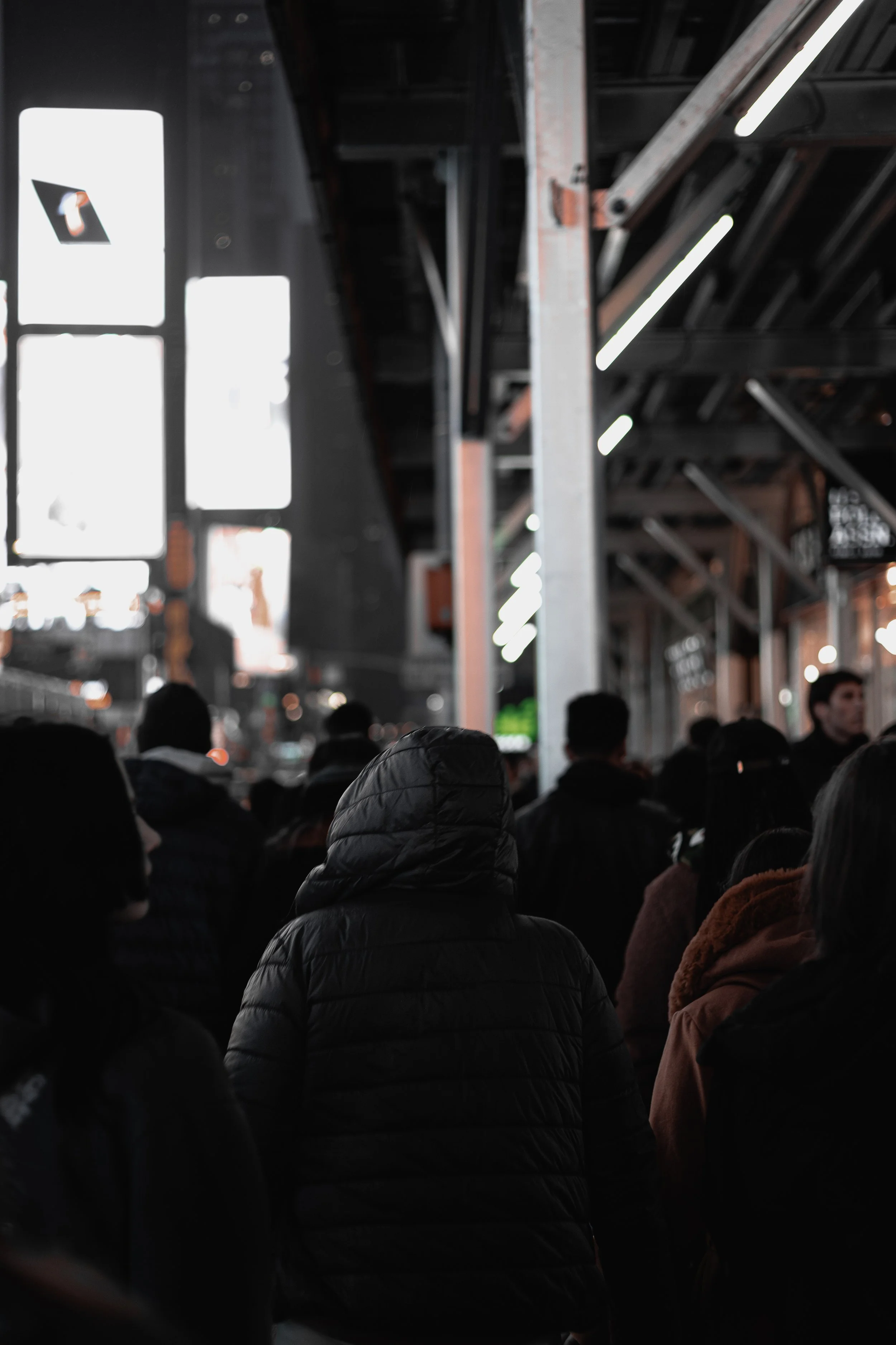 Crowd of people wearing dark jackets walking in an urban area at night with bright lights and large screens in the background.