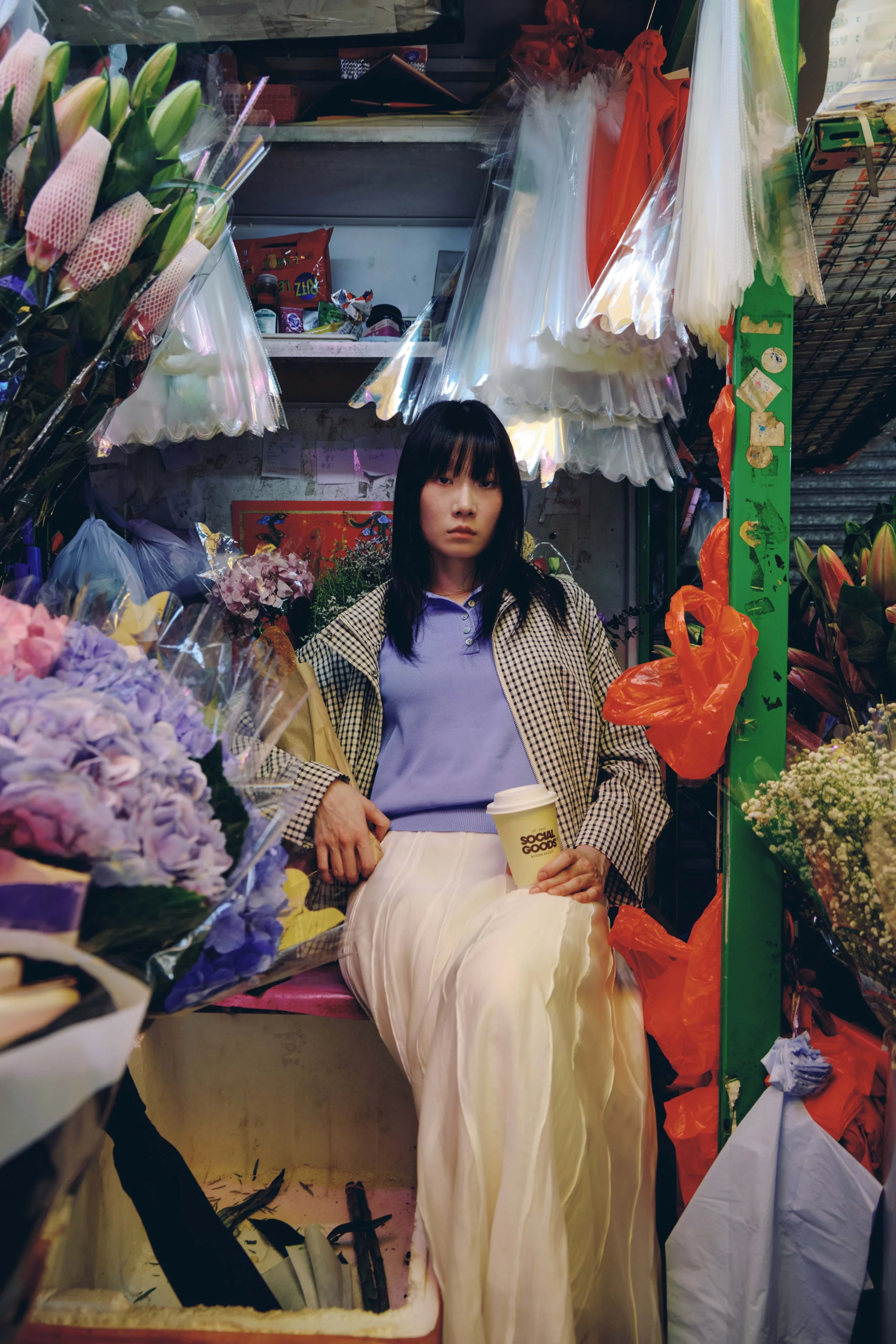 A young flower shop worker sits among vibrant blooms and floral supplies in a bustling Hong Kong flower market, holding a takeaway coffee cup.