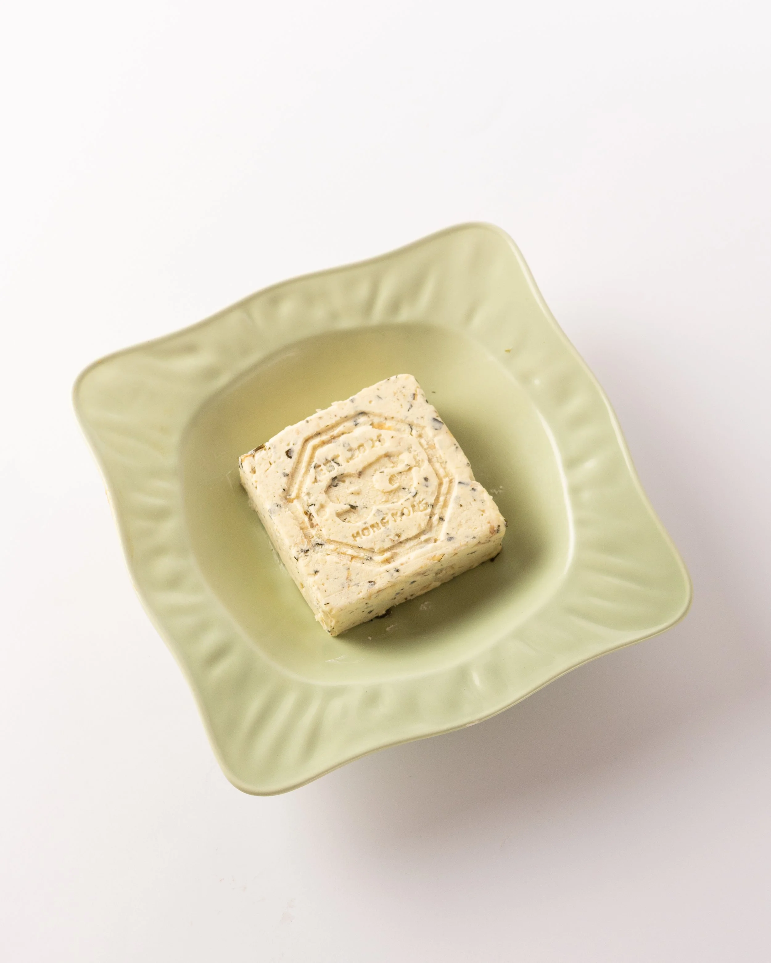 A light green ceramic dish with a square-shaped, creamy, textured dessert with visible bits of nuts or fruit on top.
