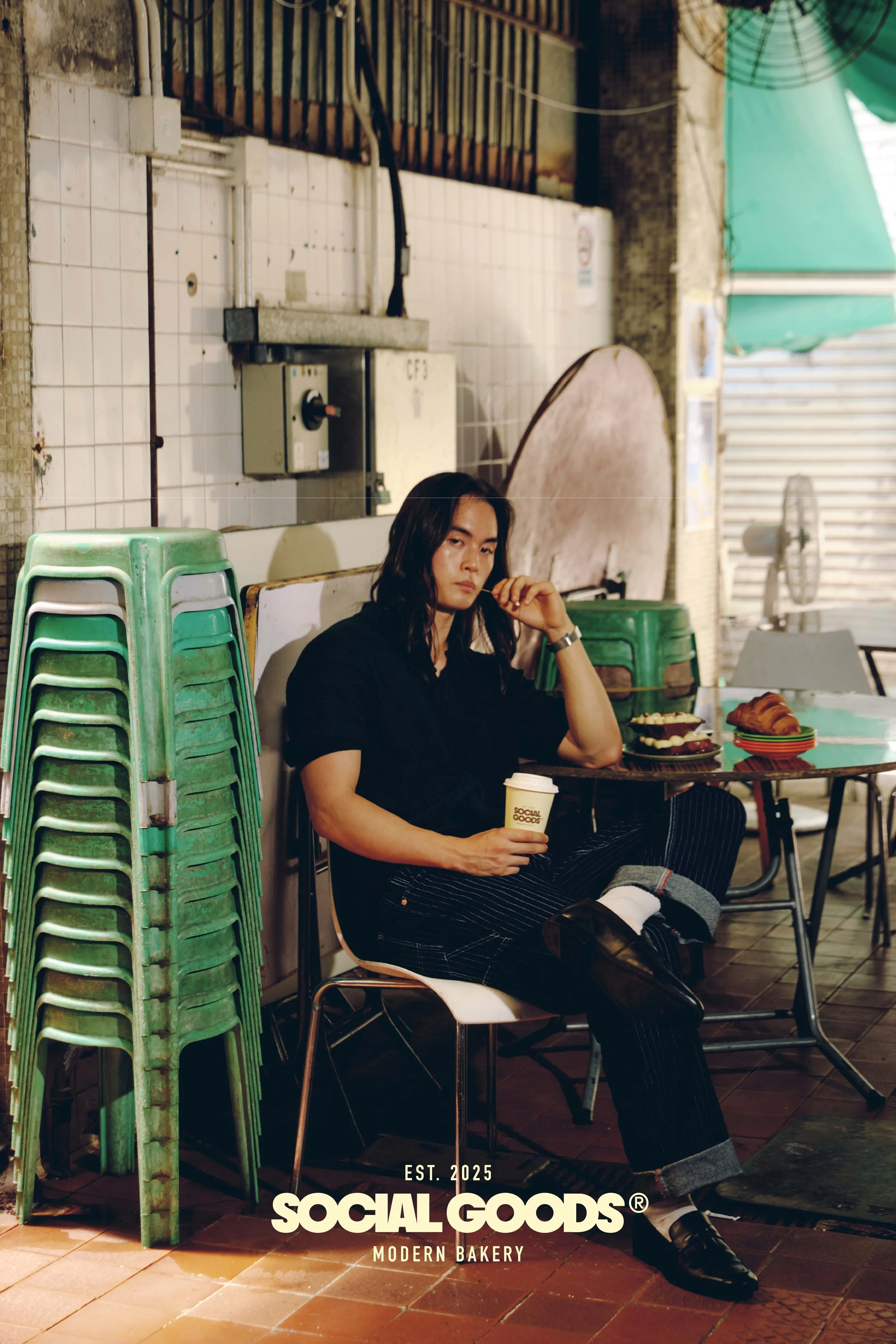 A customer sits with a Social Goods coffee cup among green chairs and pastries in the bakery's industrial-style interior.