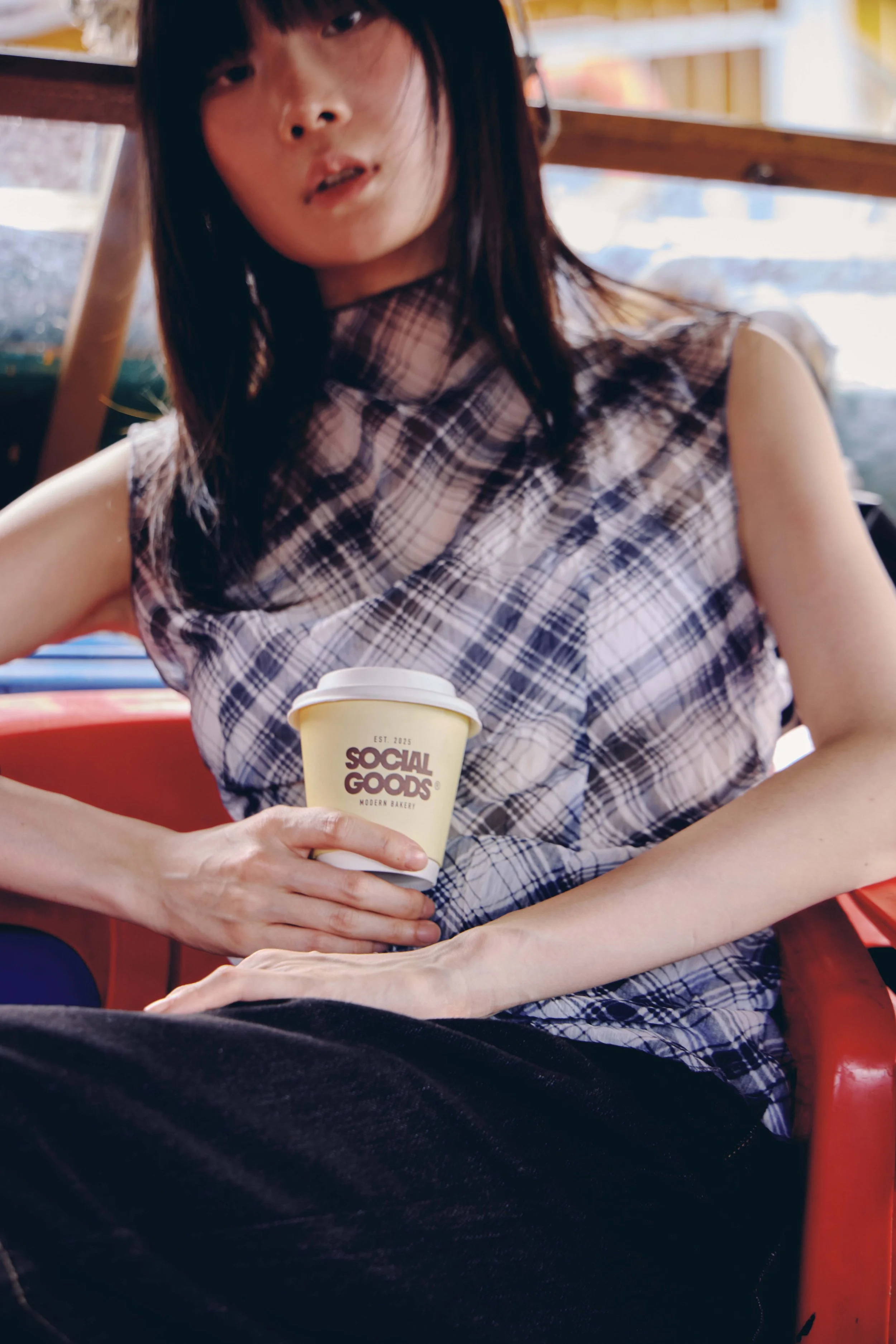 A woman sits outdoors holding a Social Goods coffee cup, with a wooden neighbourhood structure behind her.