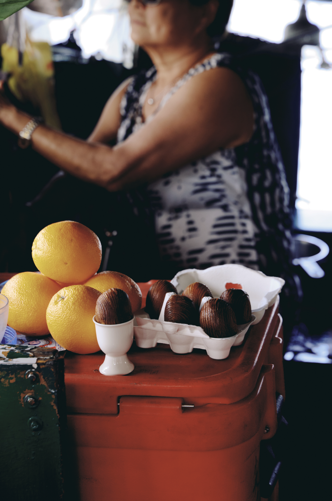 Red bucket of oranges and a grapefruit half on an outdoor market table, with a vendor in the background.