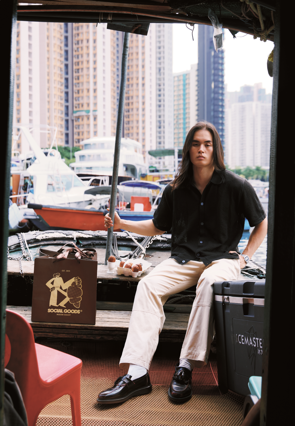 Young man sitting on a marina dock with a Social Goods shopping bag, cooler, and food tray, city skyline behind him.